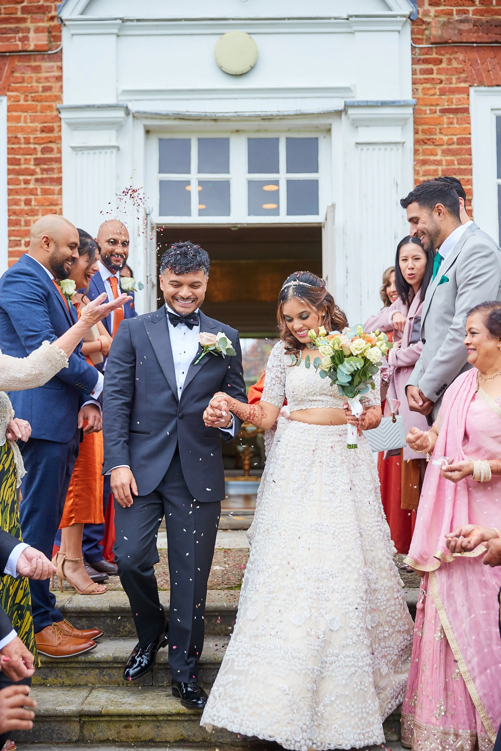 A bride and groom walking down steps after wedding, surrounded by friends and family celebrating with confetti outside of a brick building.
