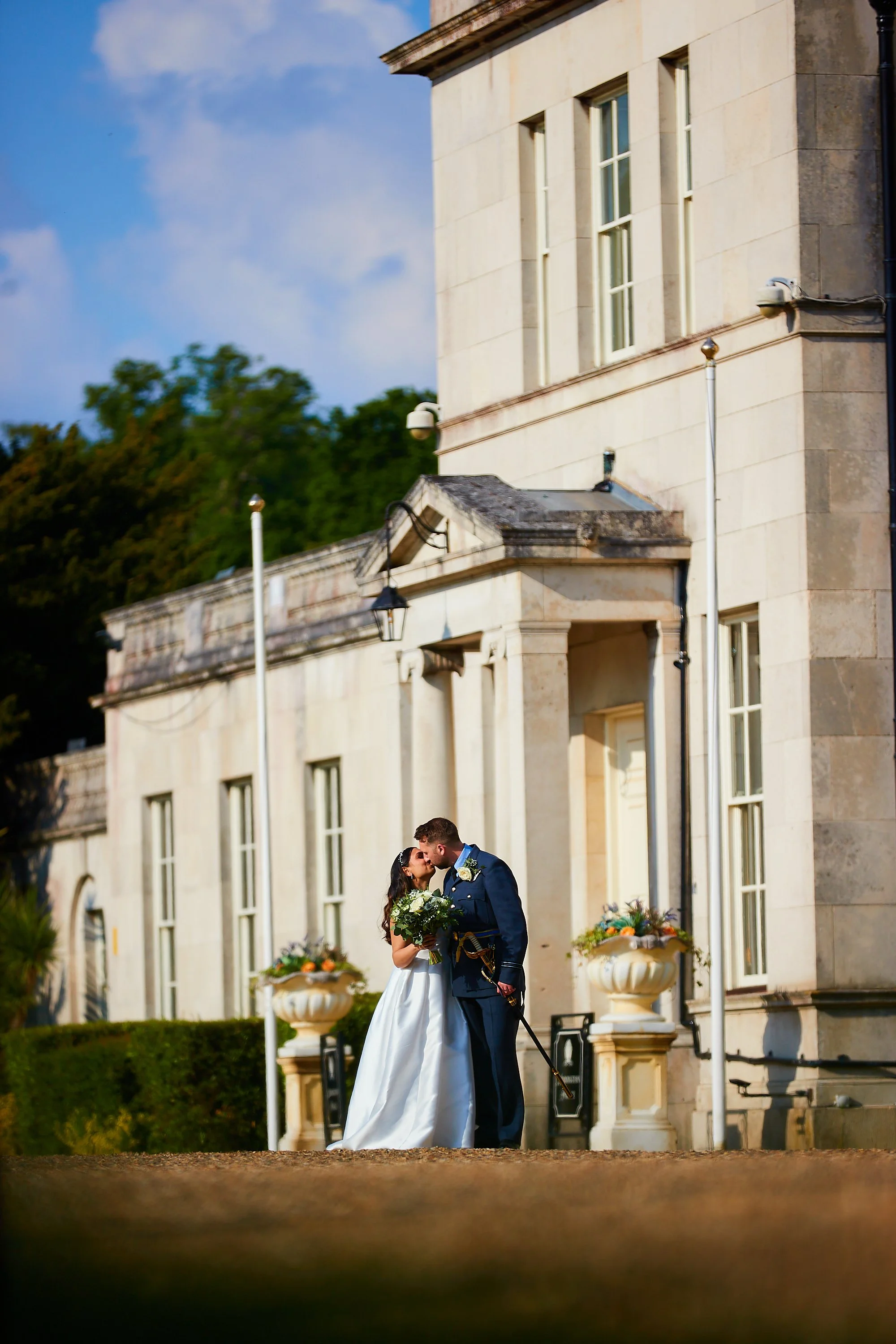 A bride and groom sharing a kiss outside a historic building, with the bride holding a bouquet of flowers, during a wedding ceremony.