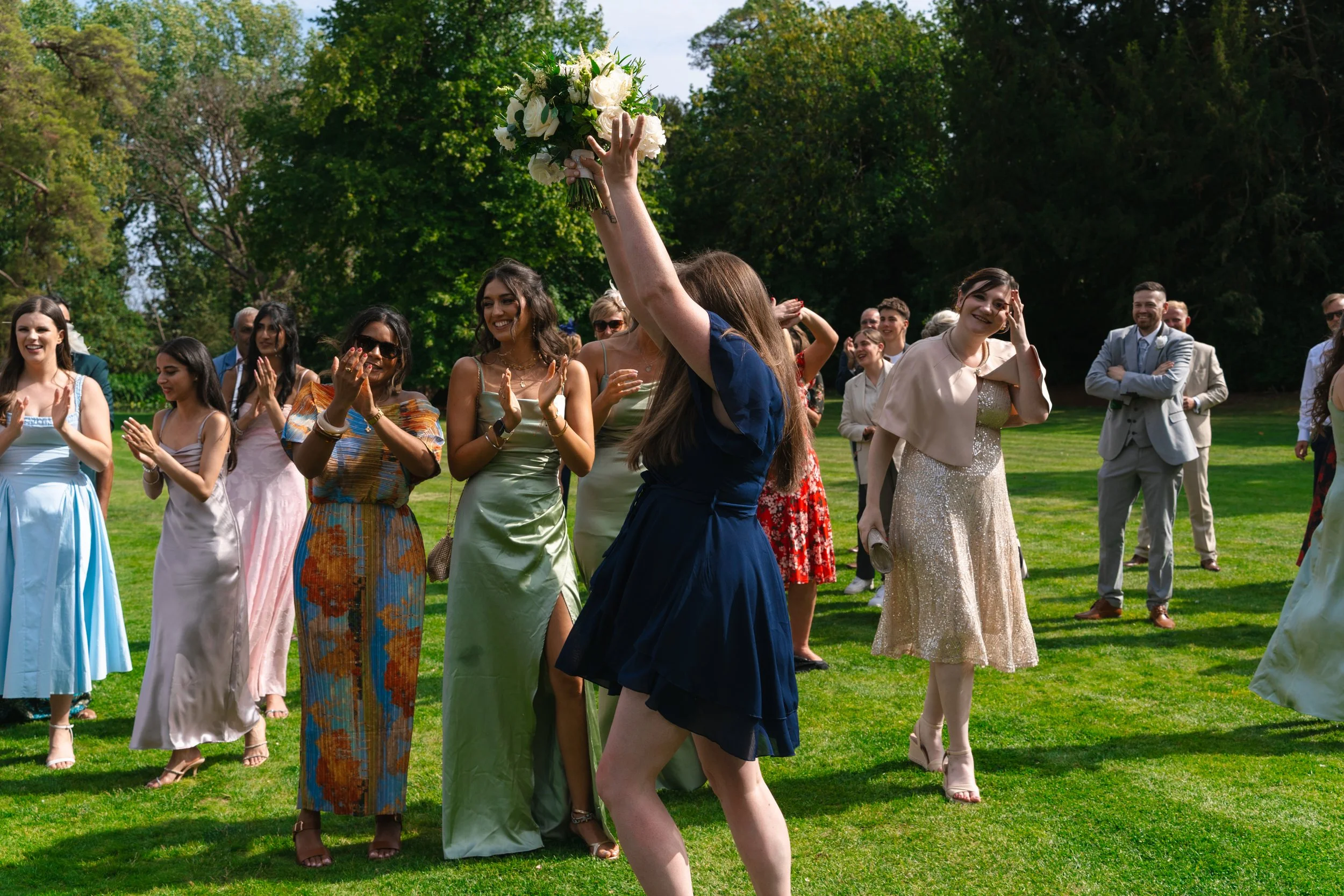 A woman in a blue dress holding a bouquet over her head, celebrating at outdoor wedding reception while another woman in a gold dress reacts with emotion. Guests in colorful dresses and suits are standing on a lush green lawn with trees in the backgr