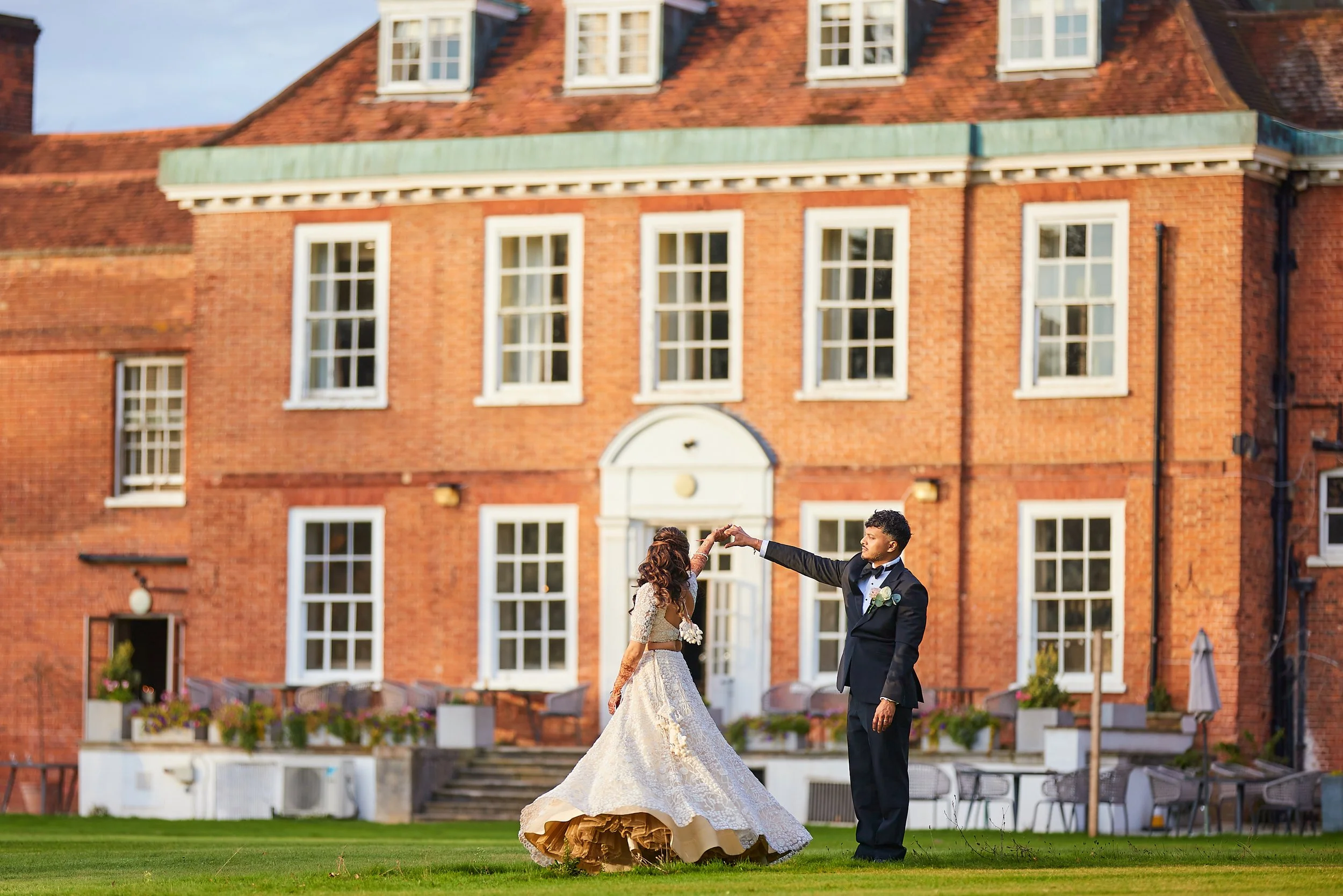 A bride and groom dance outside a large red brick house with white window frames, during sunset.