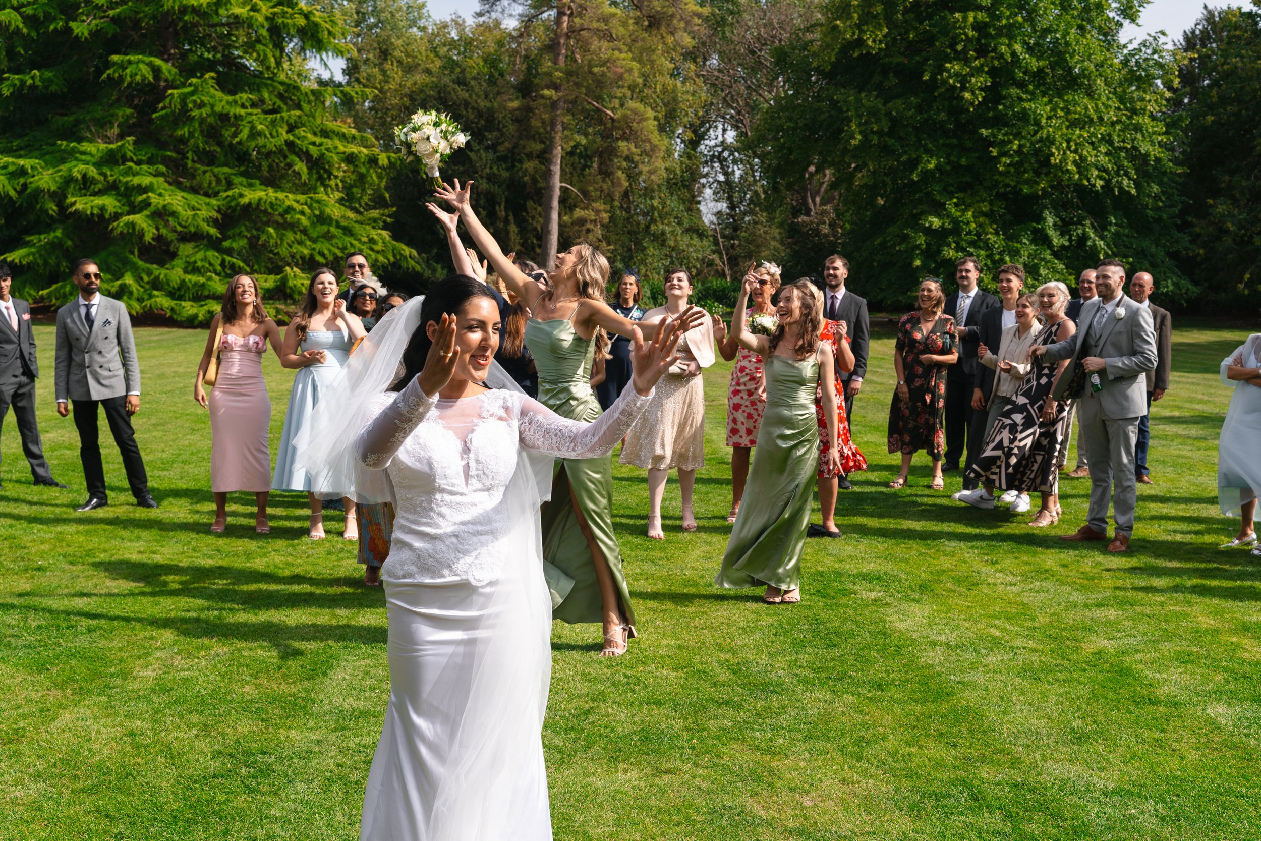 Bride throwing a bouquet of flowers with wedding guests catching it outdoors on a sunny day.