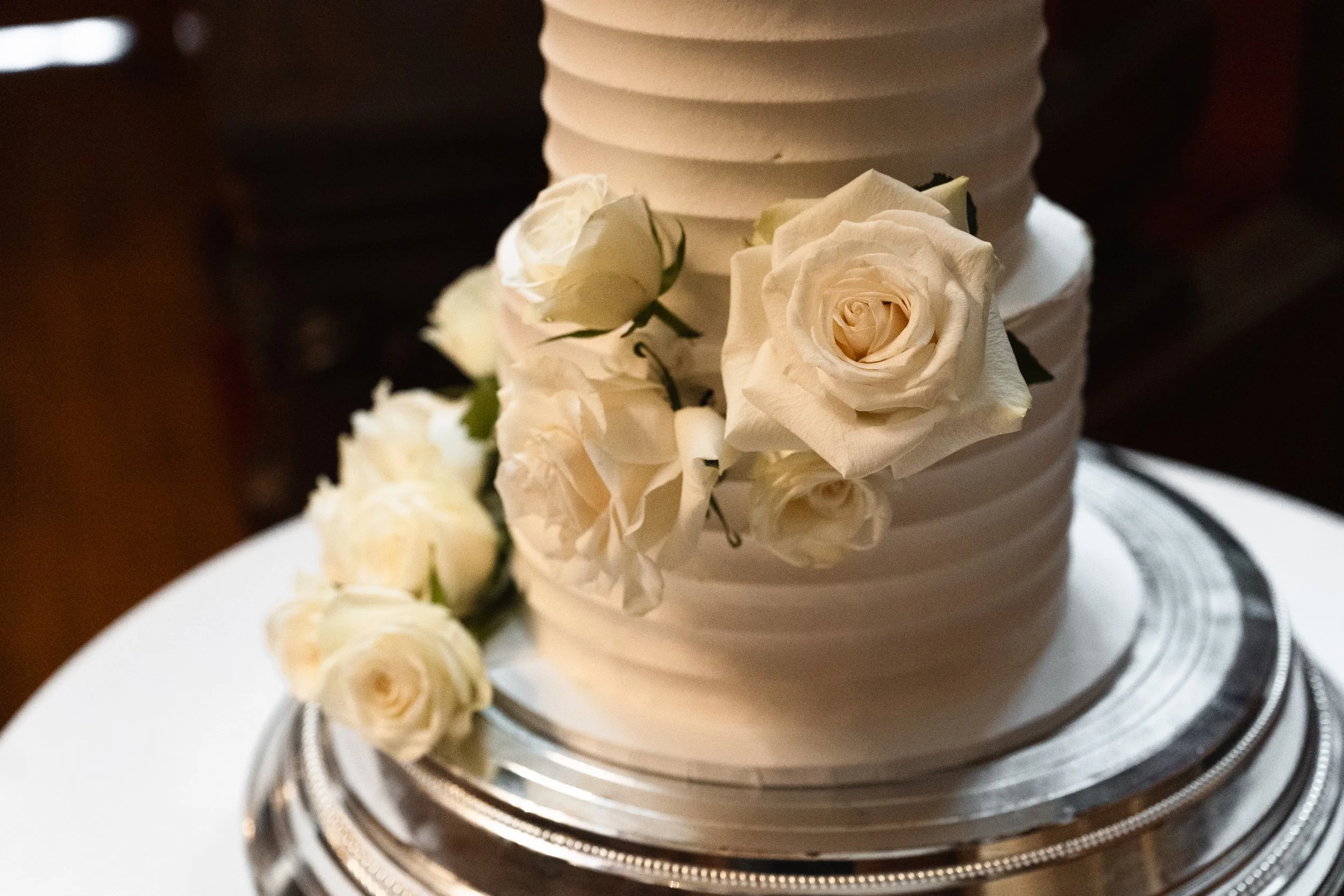 Close-up of a white wedding cake decorated with white roses placed on a silver cake stand.