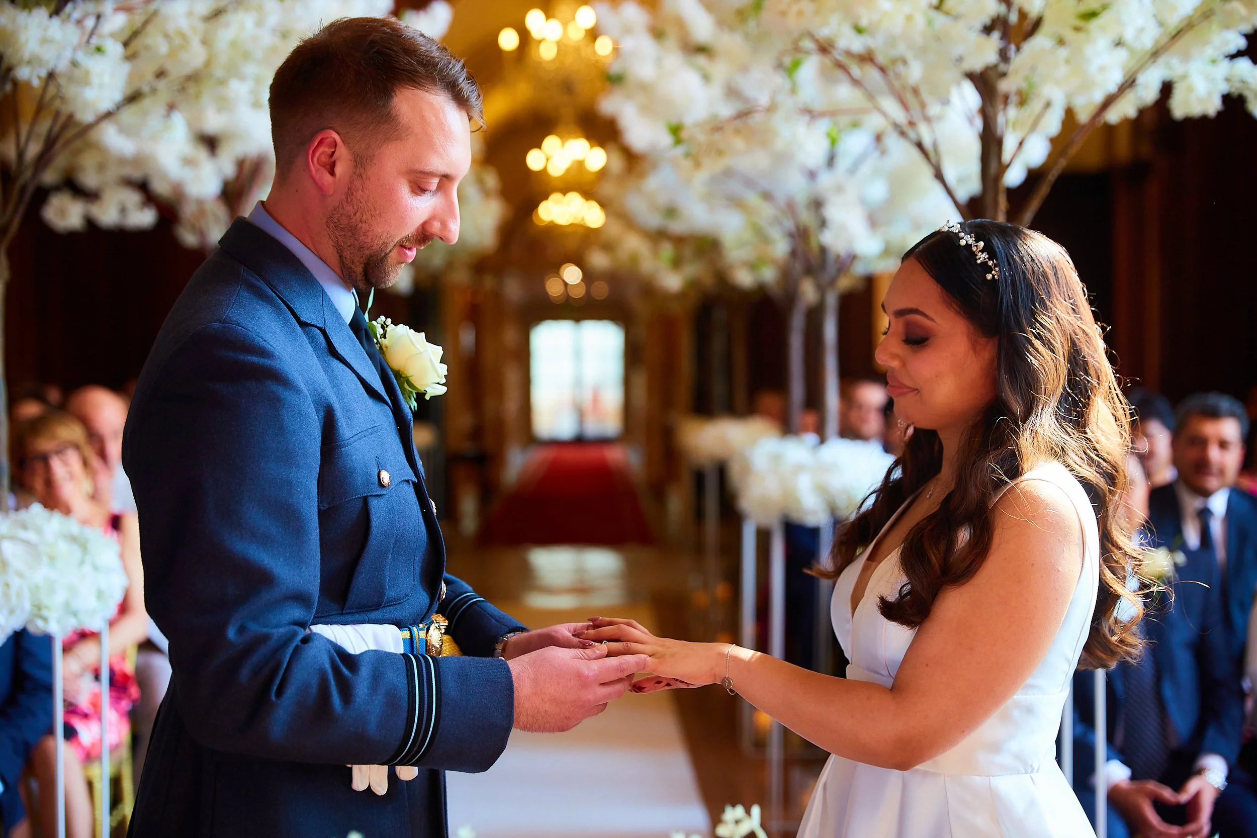 A couple getting married, the groom in a blue uniform and the bride in a white dress, exchanging rings in a decorated indoor wedding ceremony with floral arrangements and seated guests.