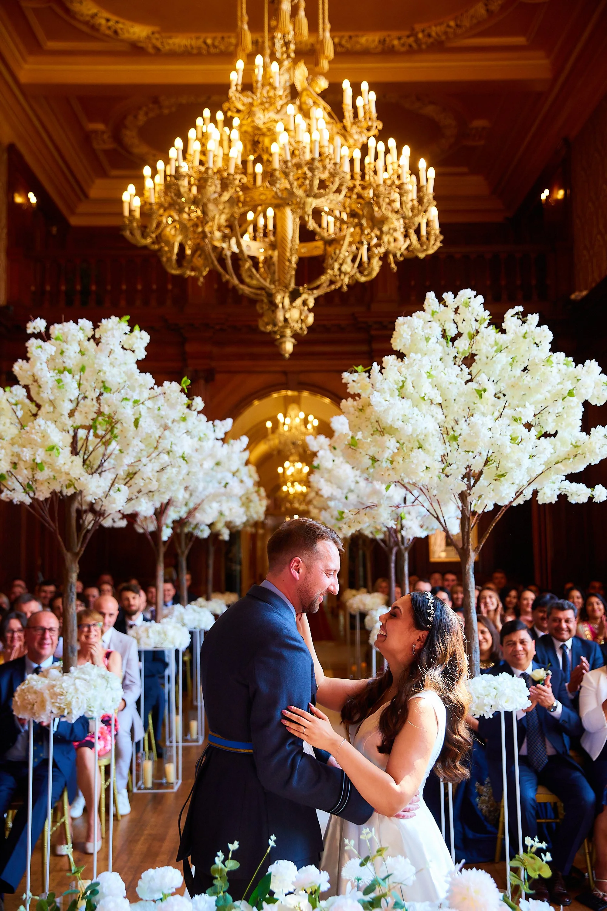 A couple gets married in a decorated indoor venue with large white floral trees, elegant chandeliers, and guests seated in the background, all joyful during the ceremony.