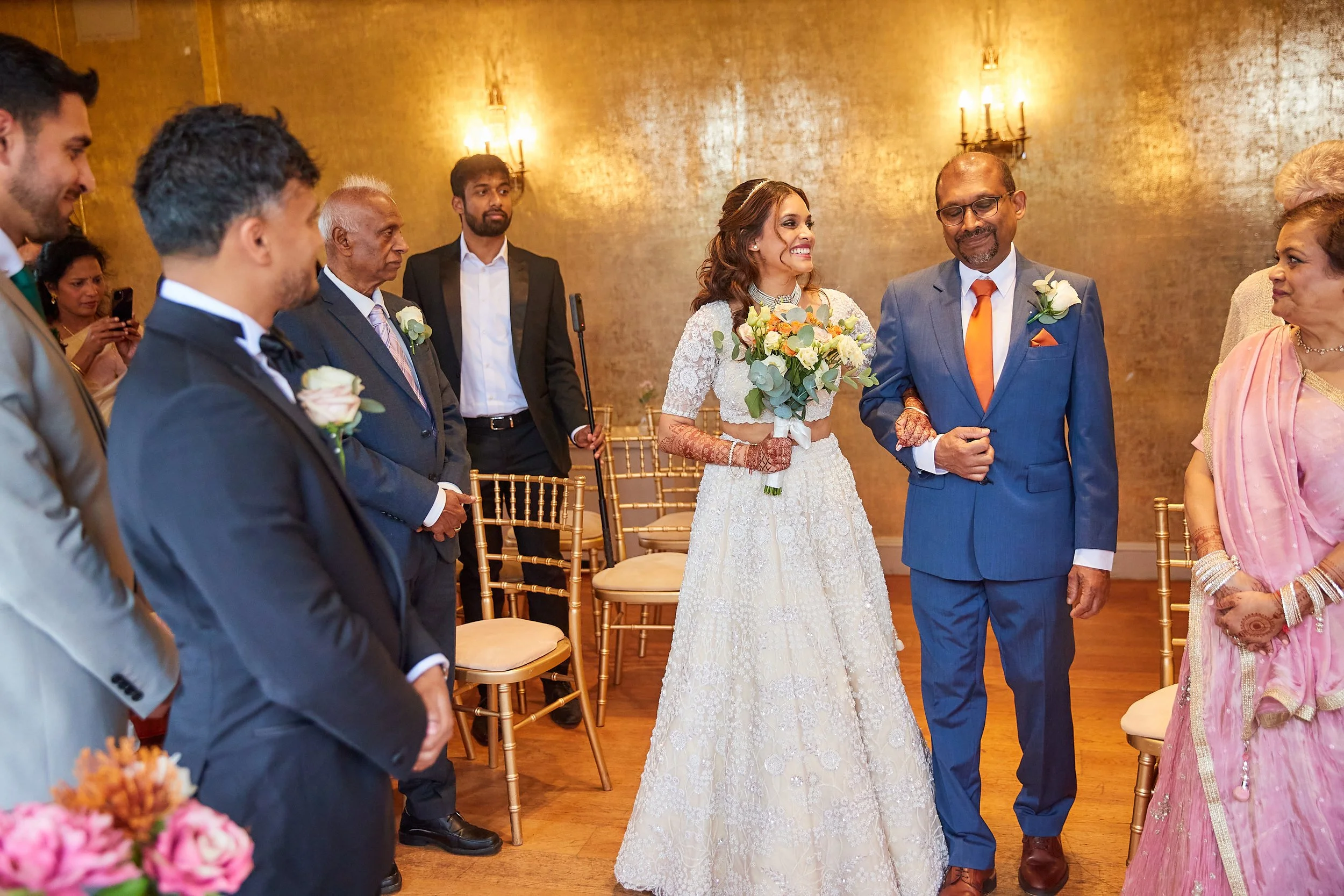 A wedding ceremony with the bride holding a bouquet, walking next to her father who is guiding her down the aisle. Guests are standing and watching. The bride is wearing a white lace dress with henna on her hands, and the father is in a blue suit wit