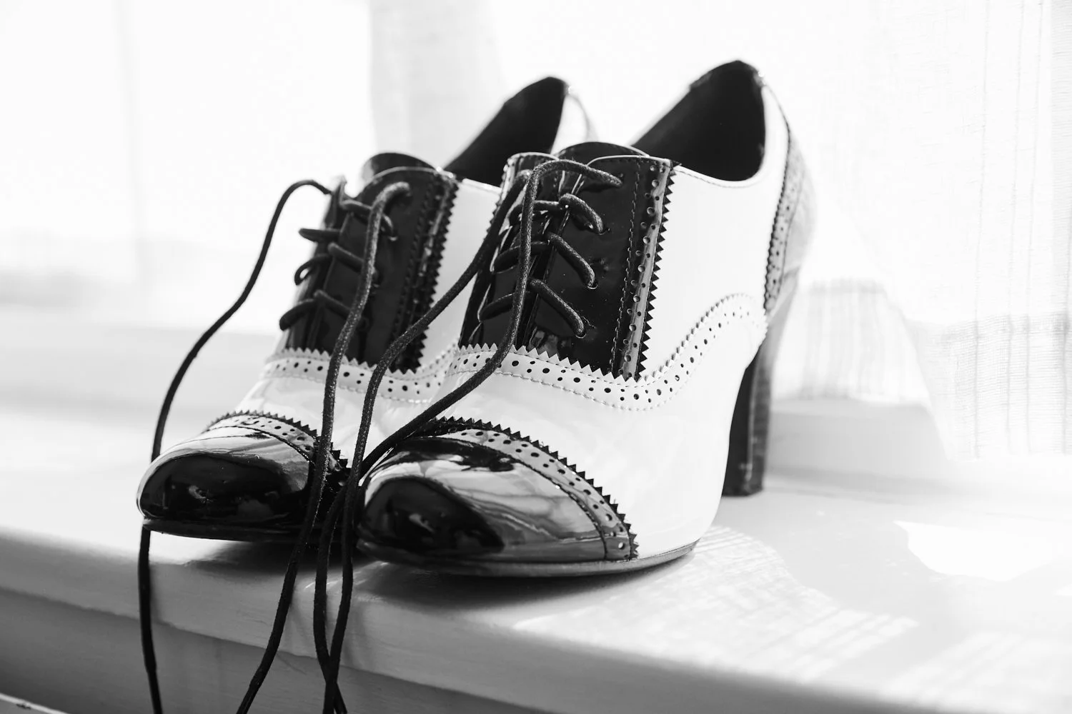 Black and white vintage-style high-heeled oxford shoes with laces, made of shiny leather, placed on a white surface near a window with sheer curtains.