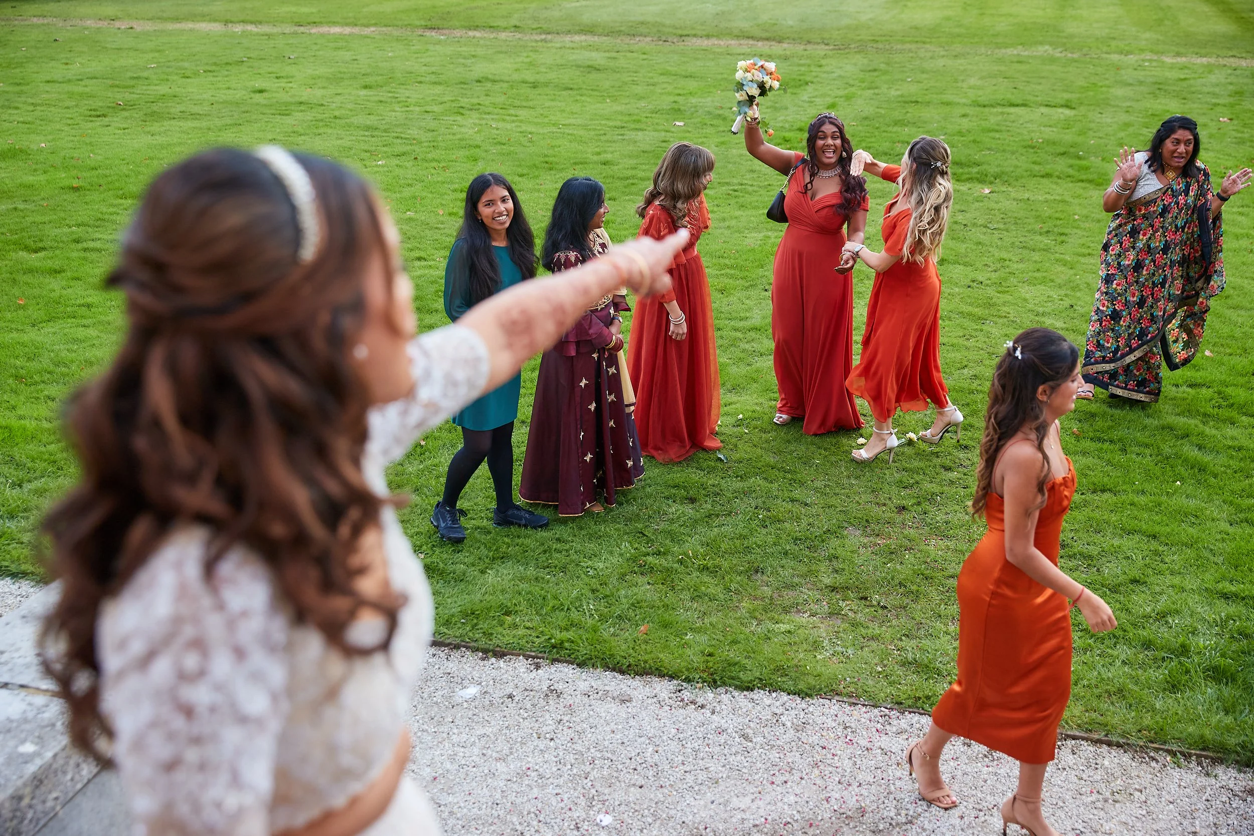 A bride in a white wedding dress points to a group of women and girls on a green lawn, celebrating a wedding. The women wear colorful dresses, and one woman holds a bouquet in the air.
