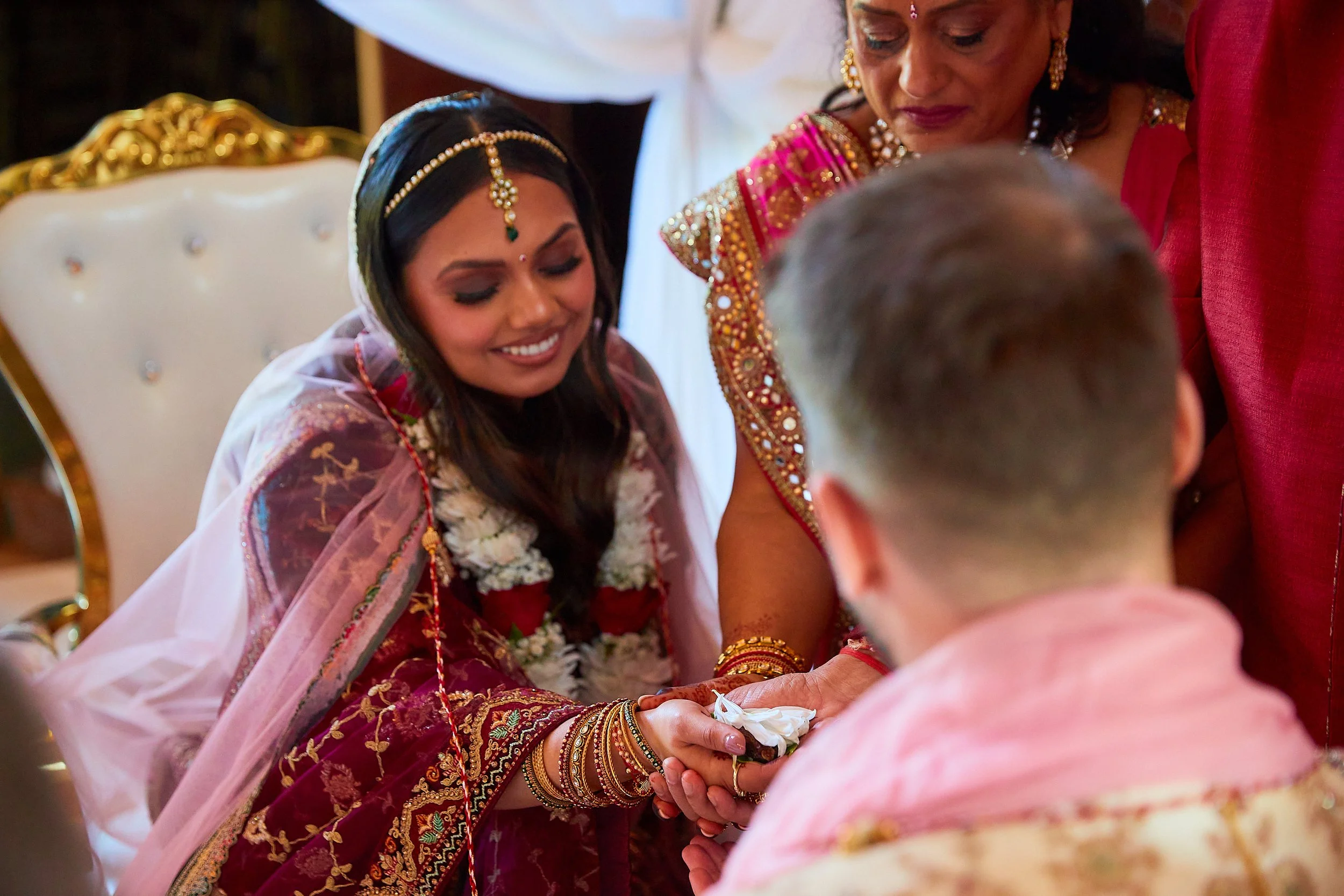 Indian bride smiling and participating in a cultural ceremony, wearing traditional attire and jewelry, engaged in a ritual with a man and woman during a wedding celebration.