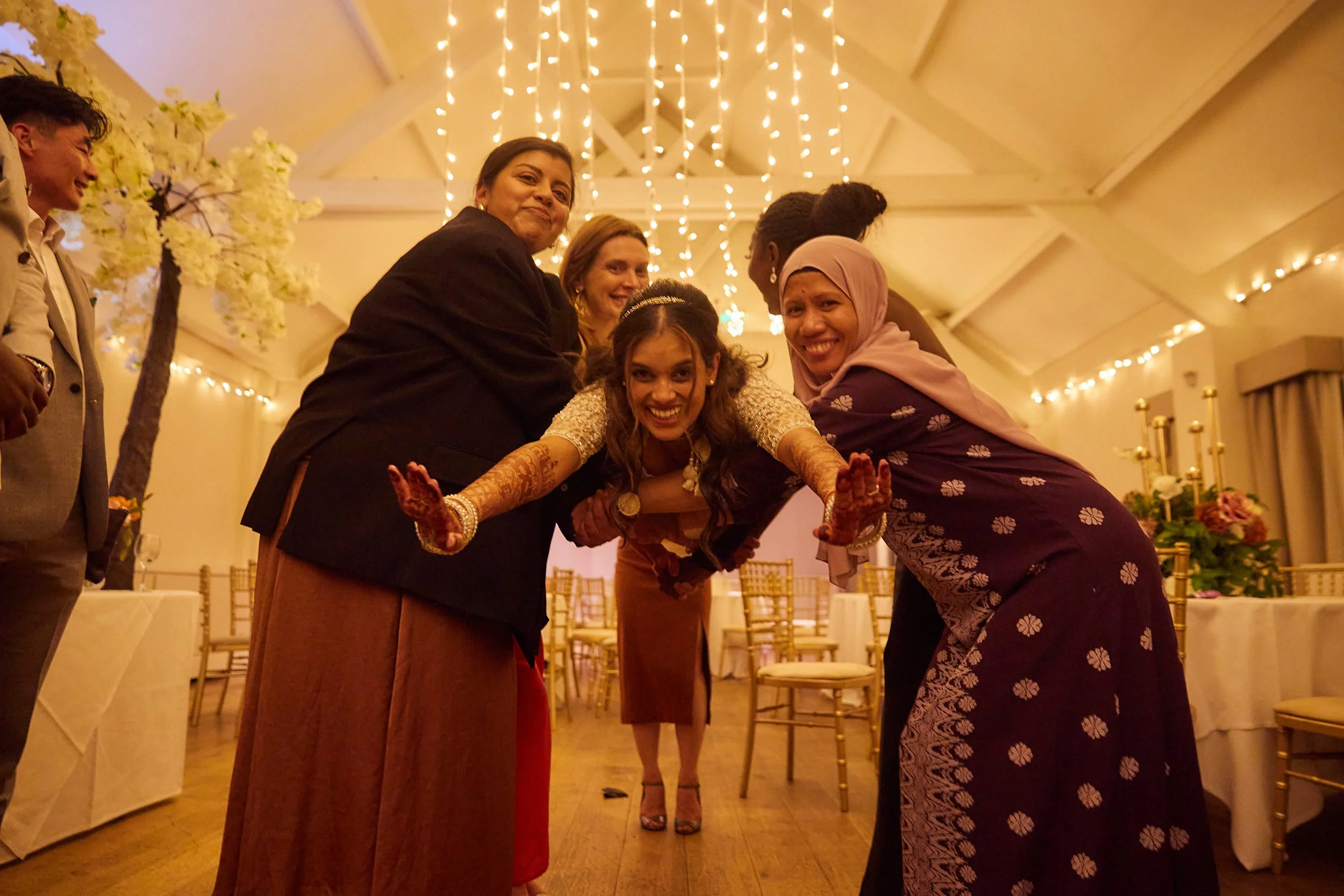 A group of women and a man at a celebration, one woman is performing an Indian dance with henna on her hands, inside a decorated hall with fairy lights and floral arrangements.