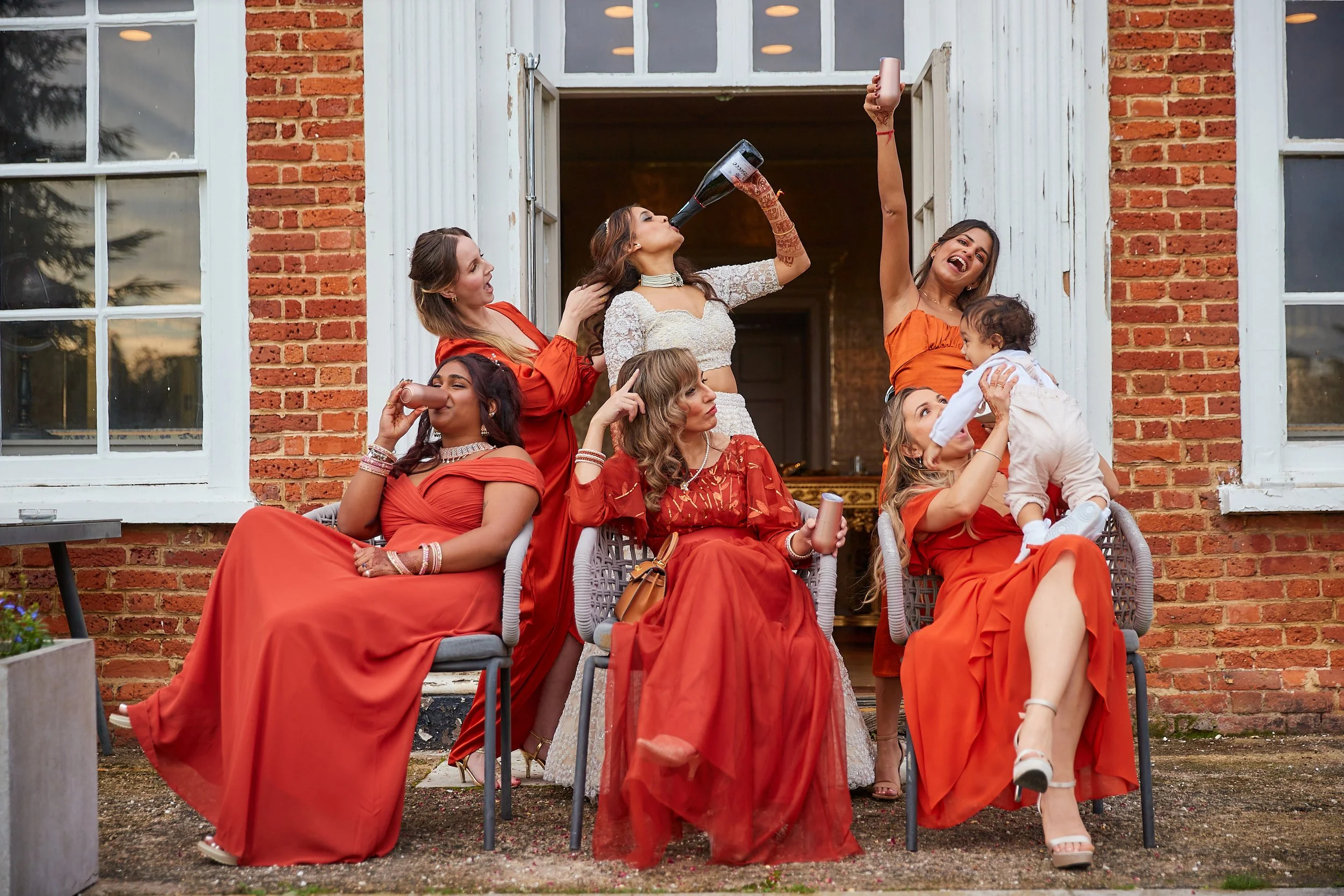 Group of women in red and white dresses celebrating outside a brick building with open window shutters. One woman is drinking from a large bottle, others are smiling, laughing, and holding drinks. A woman is holding a young child, and another woman i