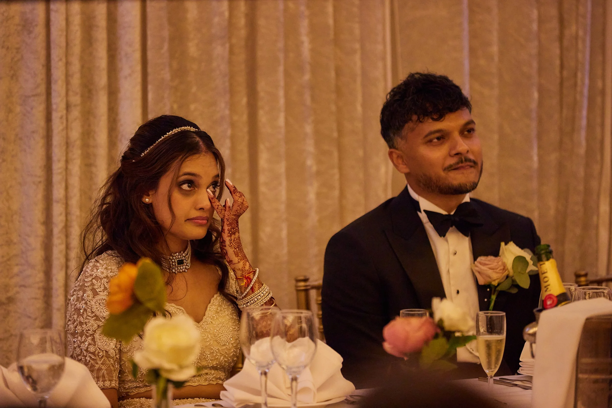 Bride and groom sitting at a wedding reception table, with the bride wiping tears and the groom looking emotional, surrounded by flowers and champagne glasses.