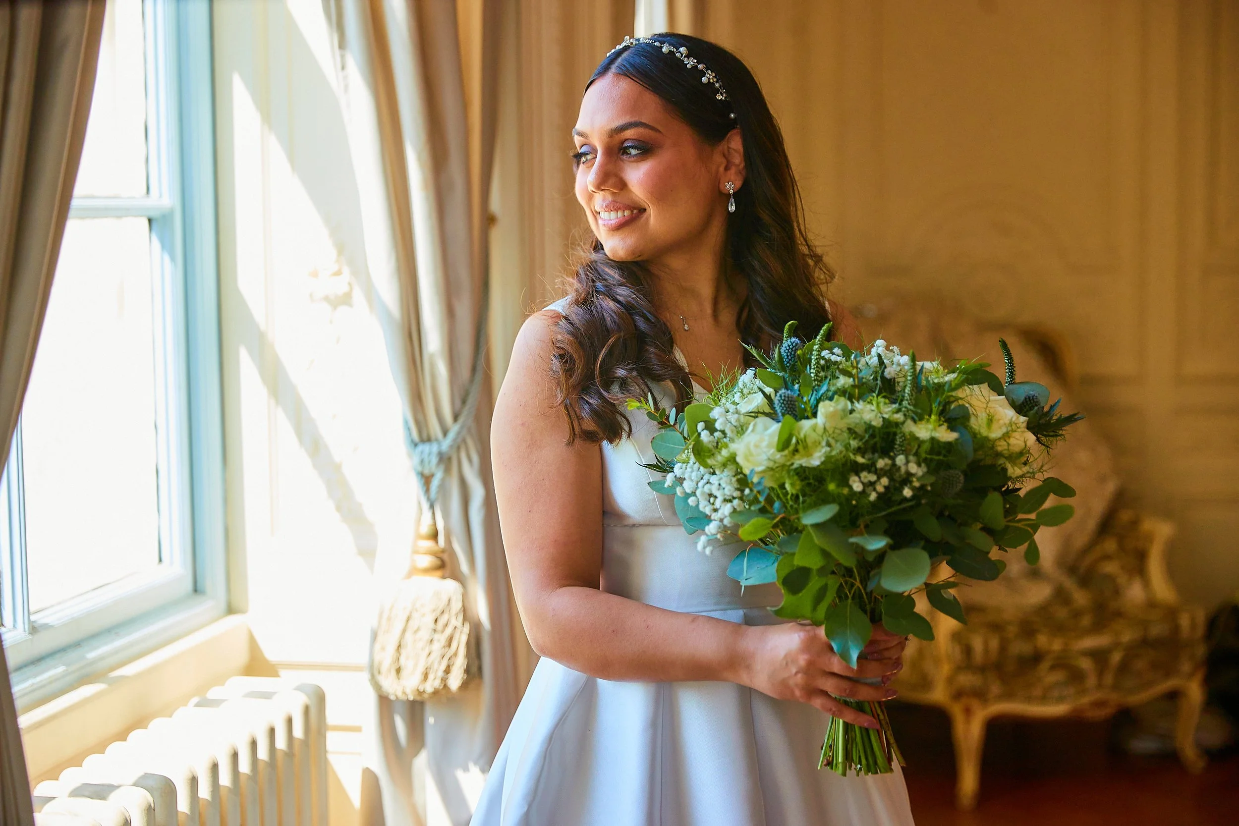 A woman in a white wedding dress holding a bouquet of white and green flowers, standing in a room with a large window and beige curtains.