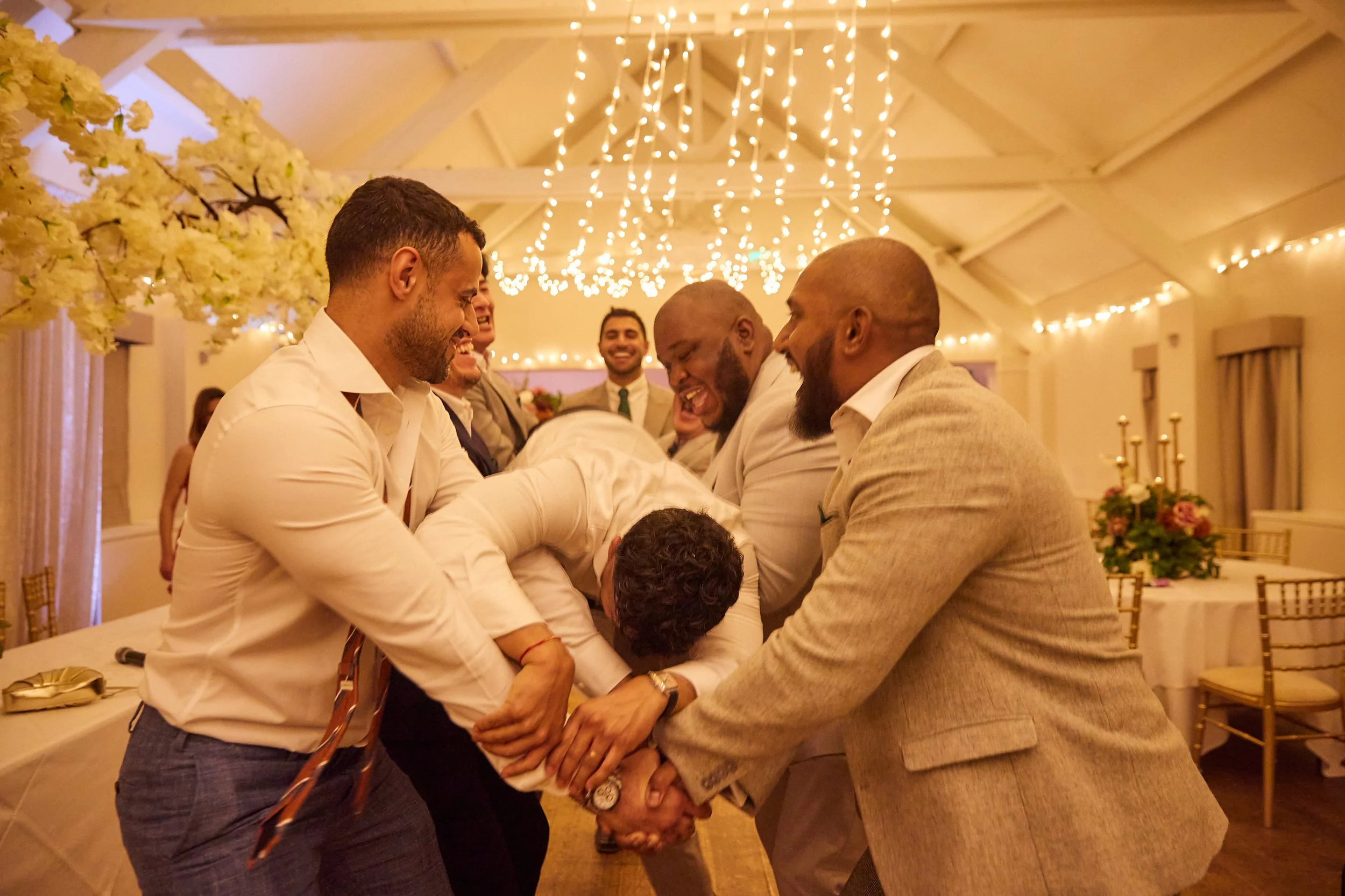 A group of men laughing and joyfully lifting a person during a celebration in an elegant decorated venue with hanging string lights and floral arrangements.