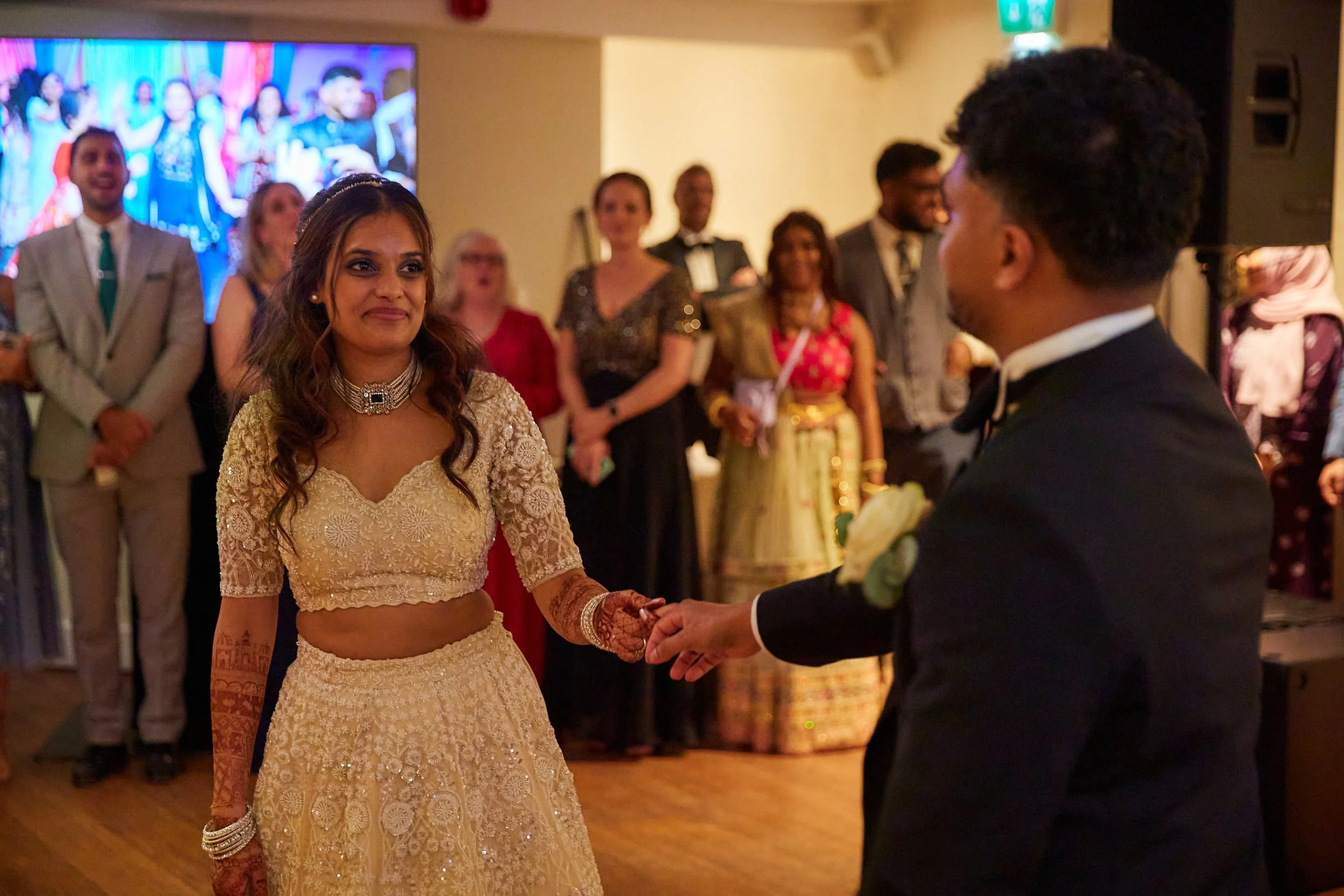 A bride and groom hold hands during their wedding dance, with guests watching in the background at a celebration.