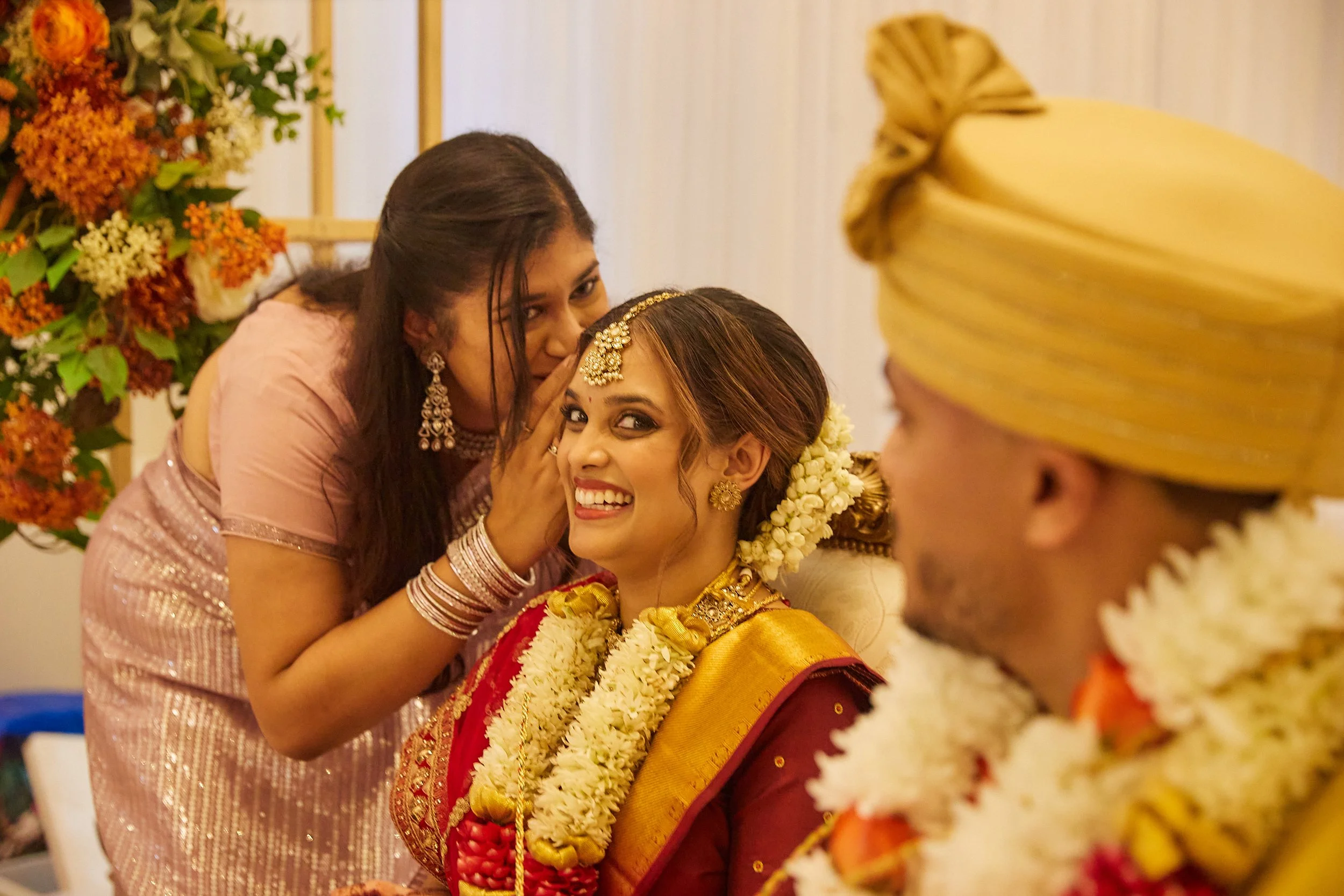 A bride smiling and a woman whispering in her ear during an Indian wedding ceremony, with a groom in traditional attire partially visible on the right. The scene is decorated with flowers.