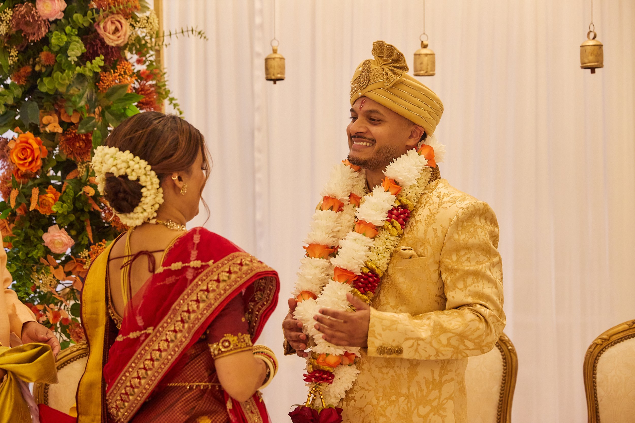 A wedding ceremony with a groom in a gold traditional outfit and turban, and a bride in a red and gold saree, exchanging vows and garlands in a decorated hall with floral arrangements and hanging lamps.