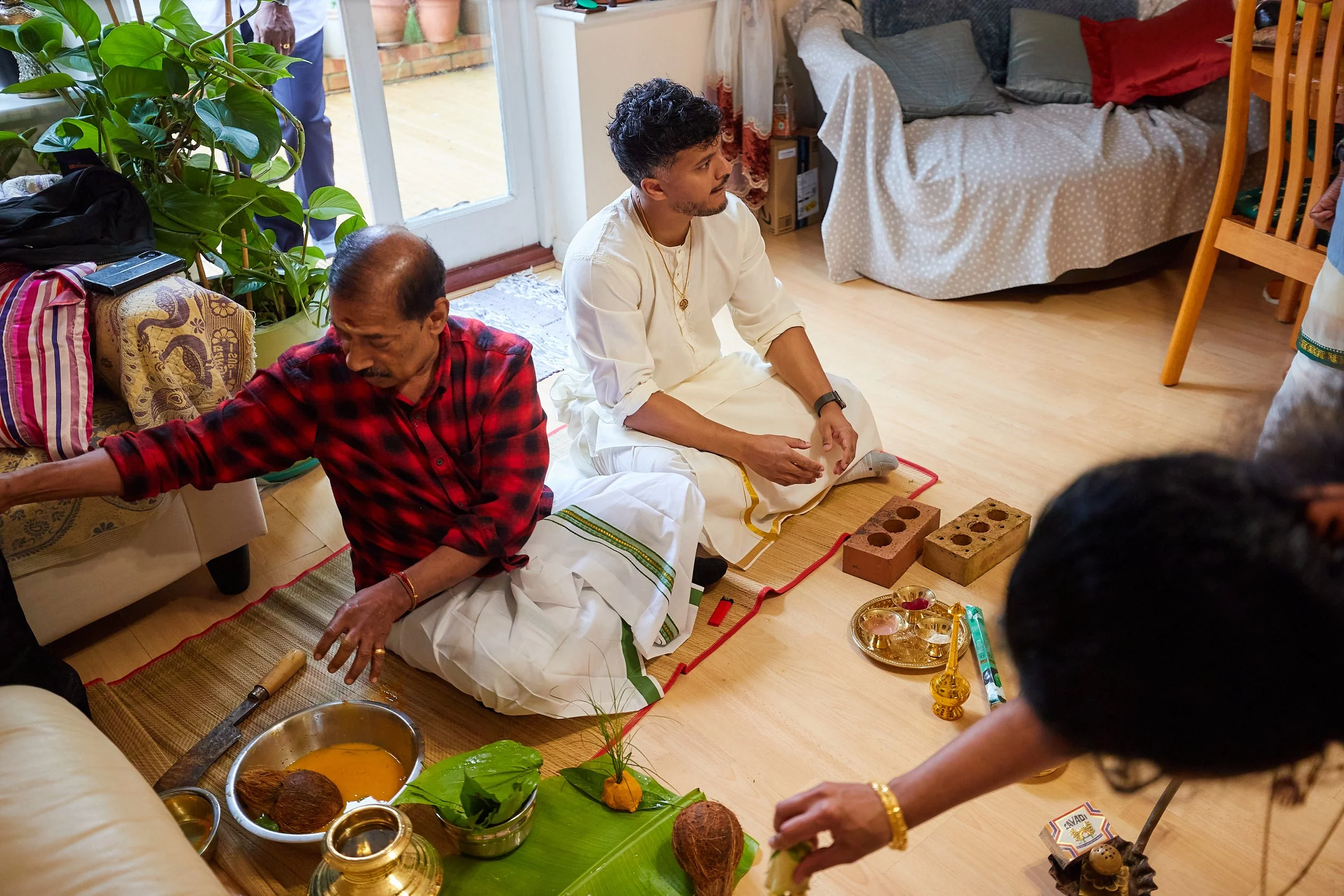 A traditional Indian pooja ceremony taking place indoors with three people participating. The elderly man in a red and black checkered shirt is reaching towards a table with religious items, while a younger man in a white traditional outfit sits on t