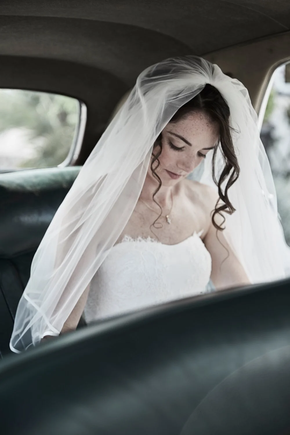 A bride with dark hair and freckles, wearing a white wedding dress and veil, sitting inside a vehicle, looking down.