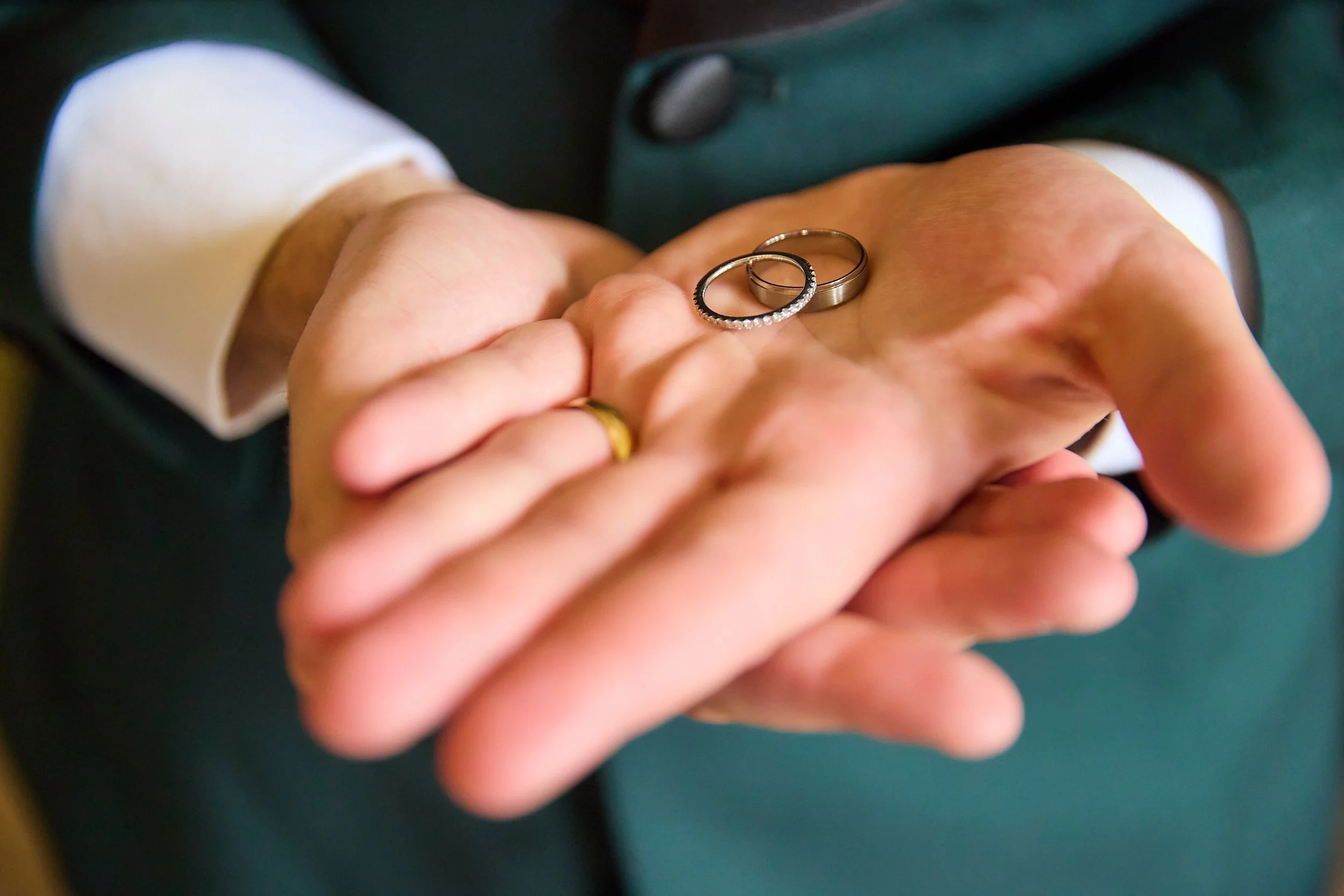 Close-up of a person wearing a white shirt and dark green jacket, holding three rings: two wedding bands and a diamond-studded ring, in their hand.