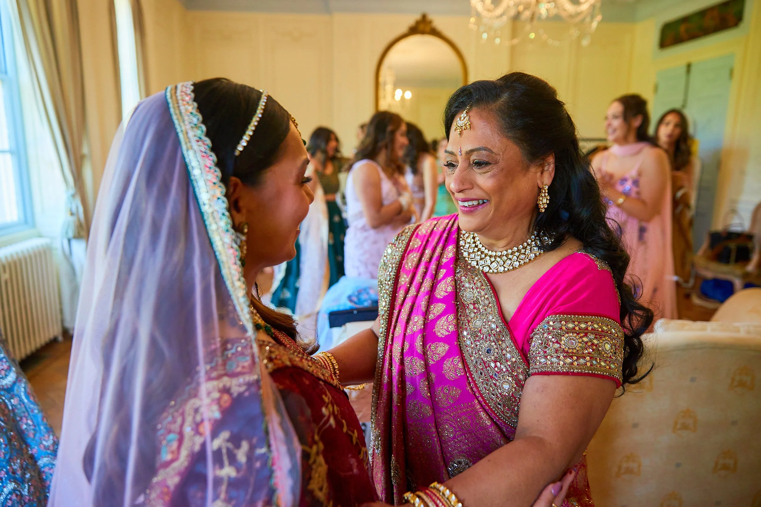Two women dressed in traditional Indian attire sharing a joyful moment, with others in the background inside a decorated room.