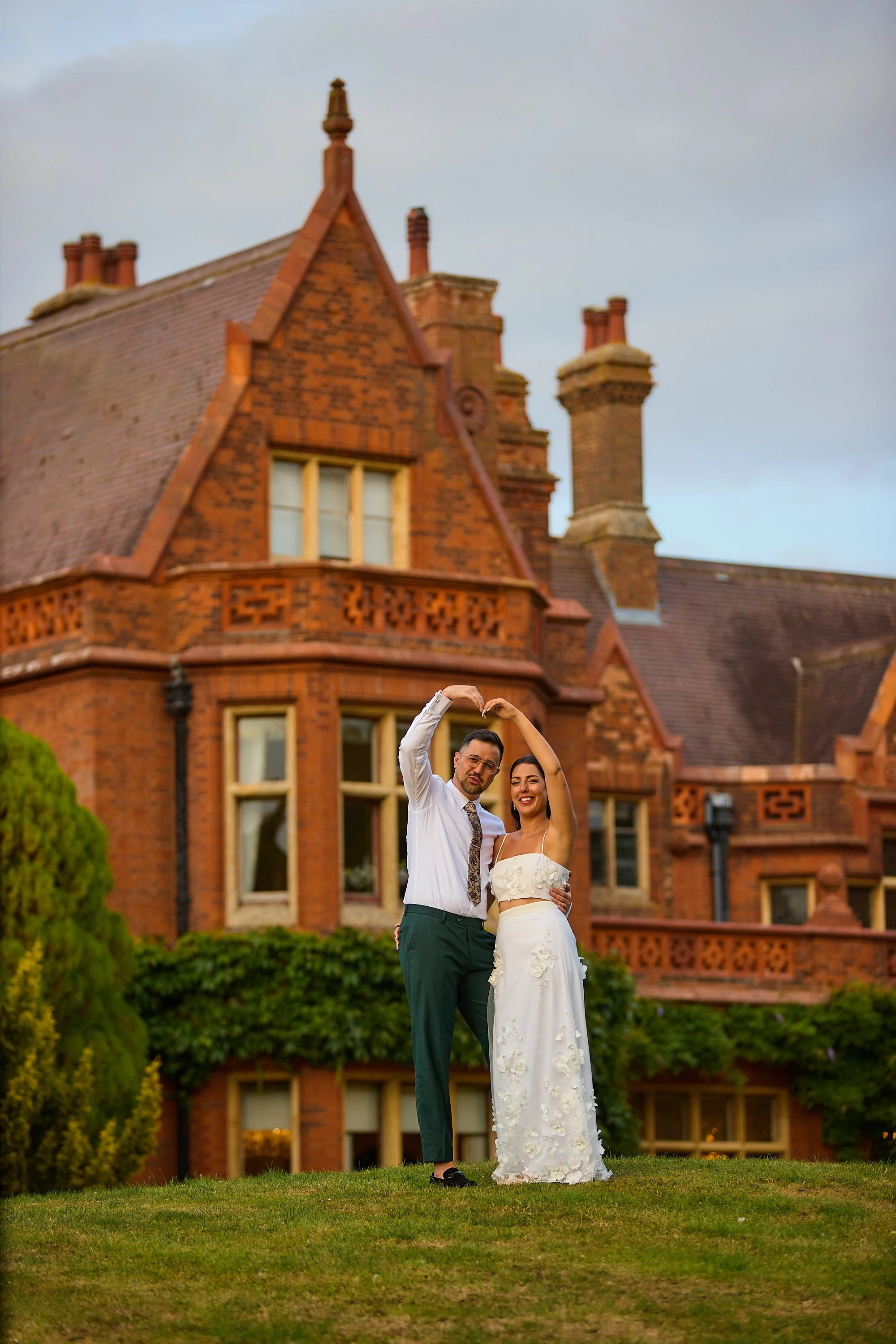 A happy couple in wedding attire dancing outdoors on a grassy area in front of a large historic red brick house with Victorian architecture.