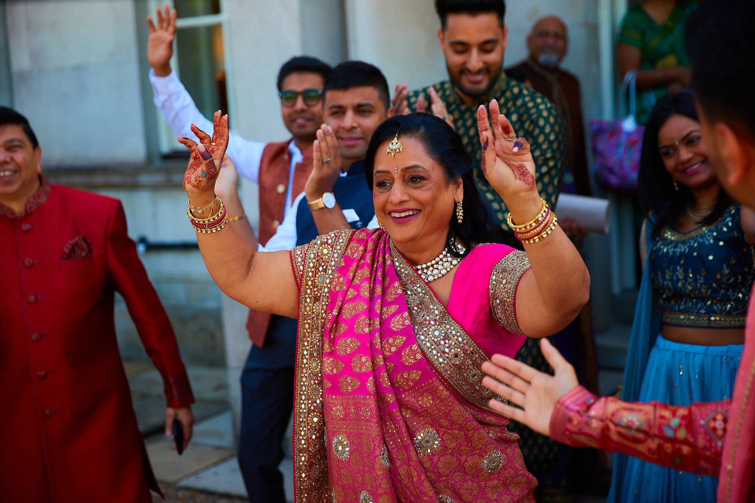 A woman in traditional Indian attire, wearing a pink saree with gold embroidery and jewelry, is smiling and raising her hands during a celebration. Several other people around her are clapping and smiling.