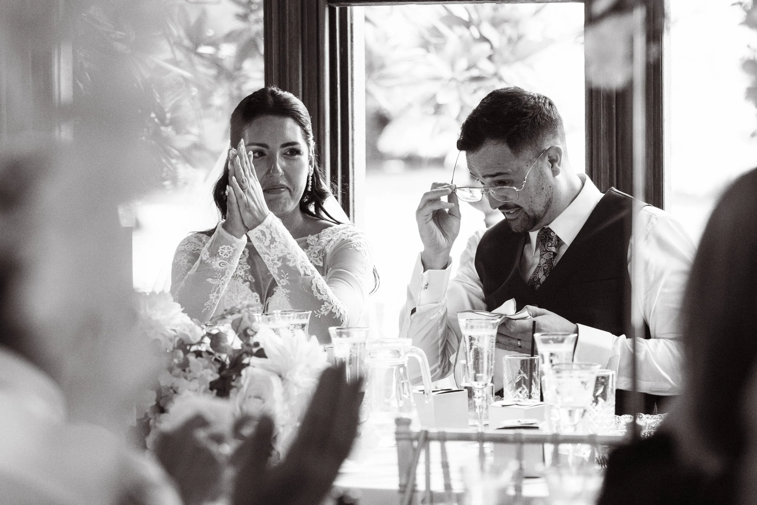 A wedding reception scene with a bride and groom sitting at a table, surrounded by glasses and a floral centerpiece. The bride appears emotional, with her hands clasped near her face, while the groom adjusts his glasses, reading a note or speech. The