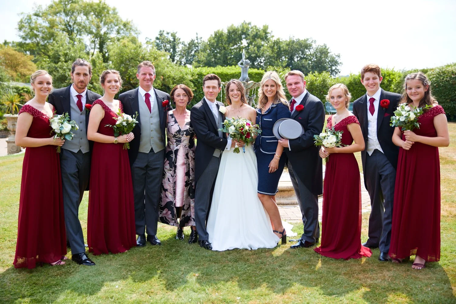 Group of people at a wedding, including the bride and groom, bridesmaids in red dresses, and groomsmen in dark suits, standing outdoors in front of a fountain and greenery.