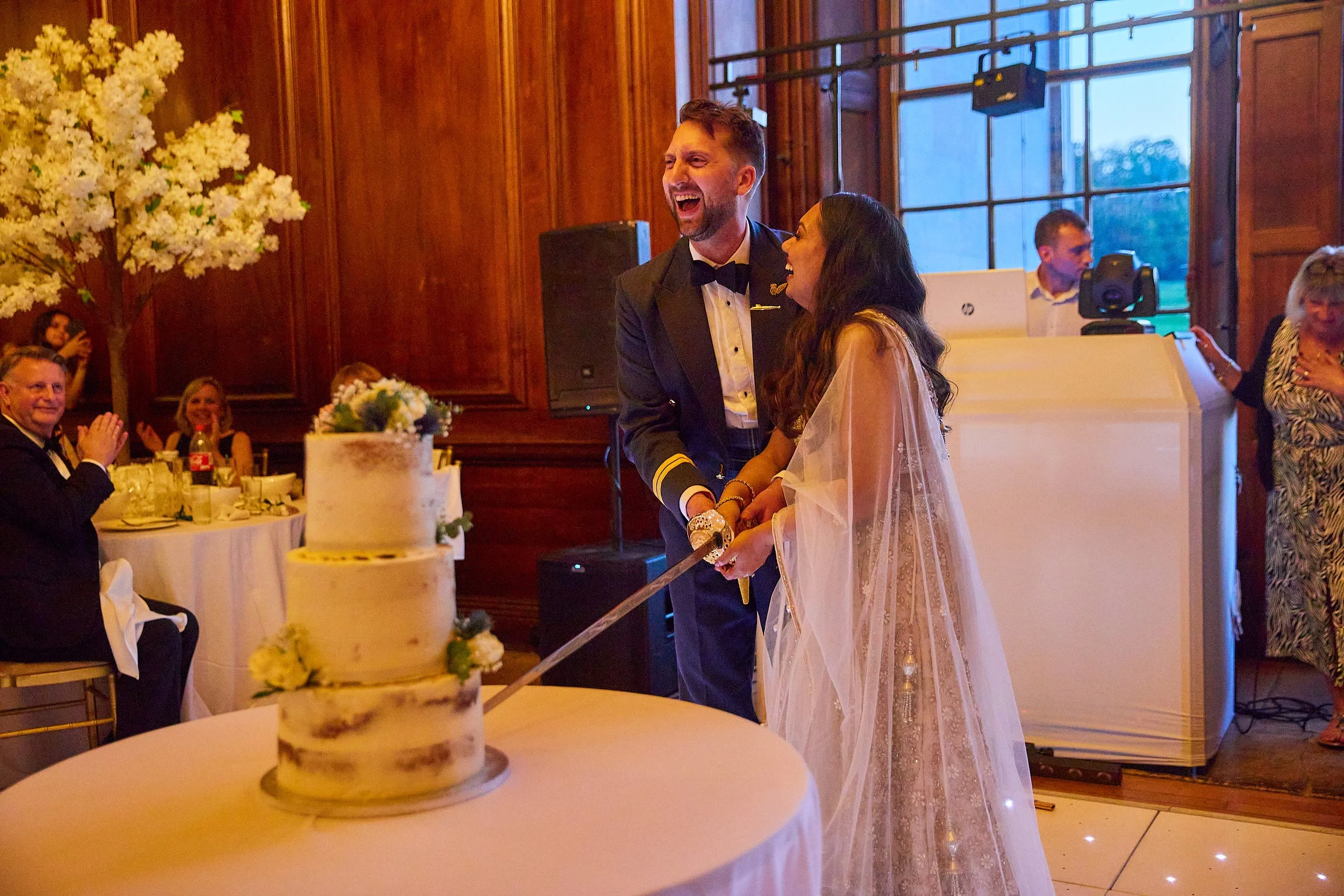 A newlywed couple celebrating their wedding with a cake-cutting ceremony. The groom is in a formal tuxedo, and the bride is wearing a wedding gown with a sheer veil. They are smiling and holding a large knife together. In the background, guests are s
