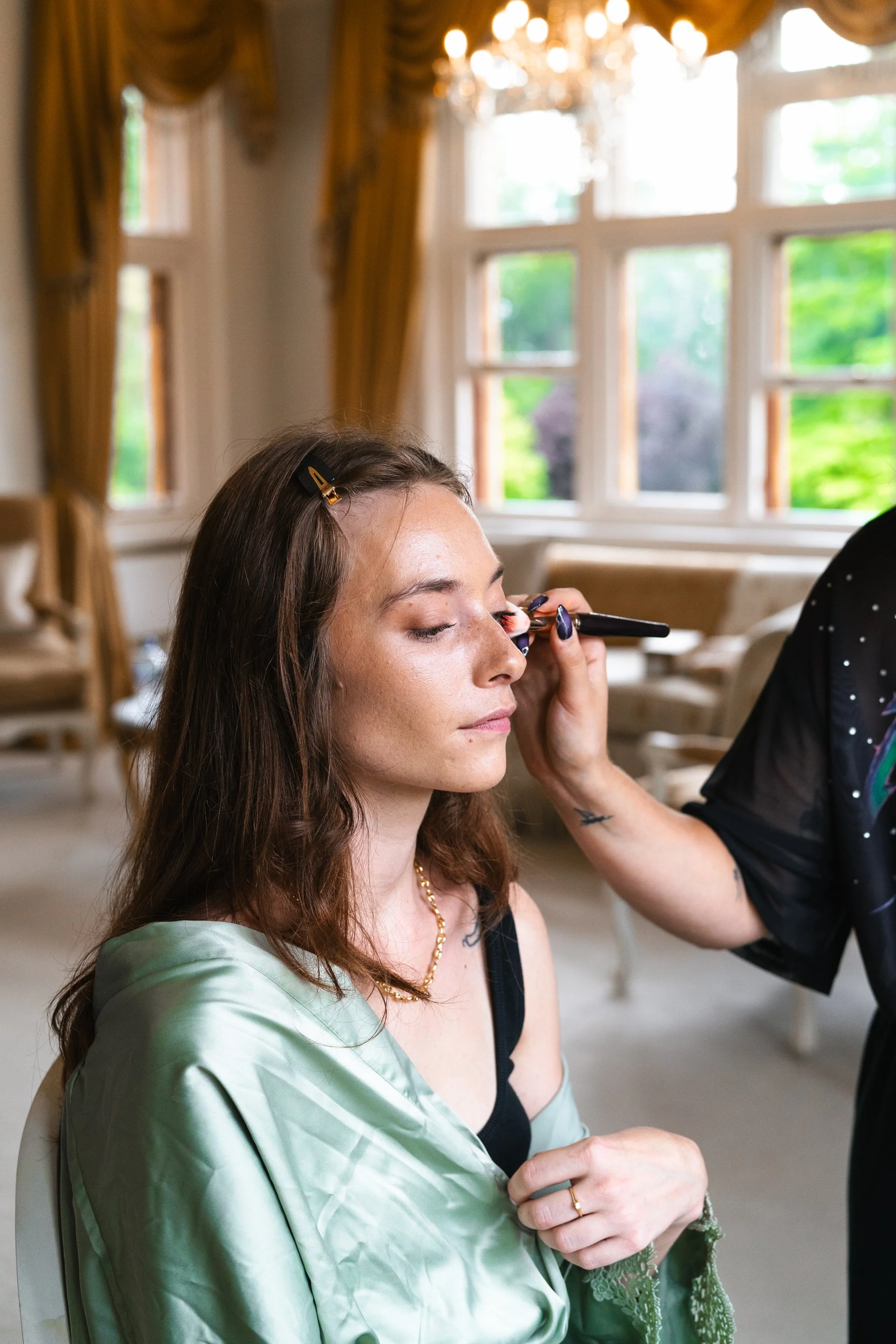 A woman with long brown hair getting her makeup done by a makeup artist in a bright, elegant room with large windows and gold curtains.