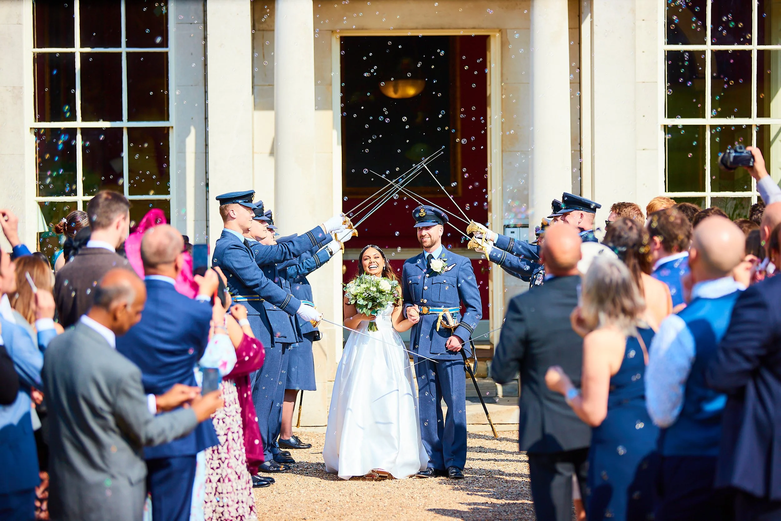 A newly married couple is standing outside a grand building while wedding guests celebrate with balloons and bubbles.