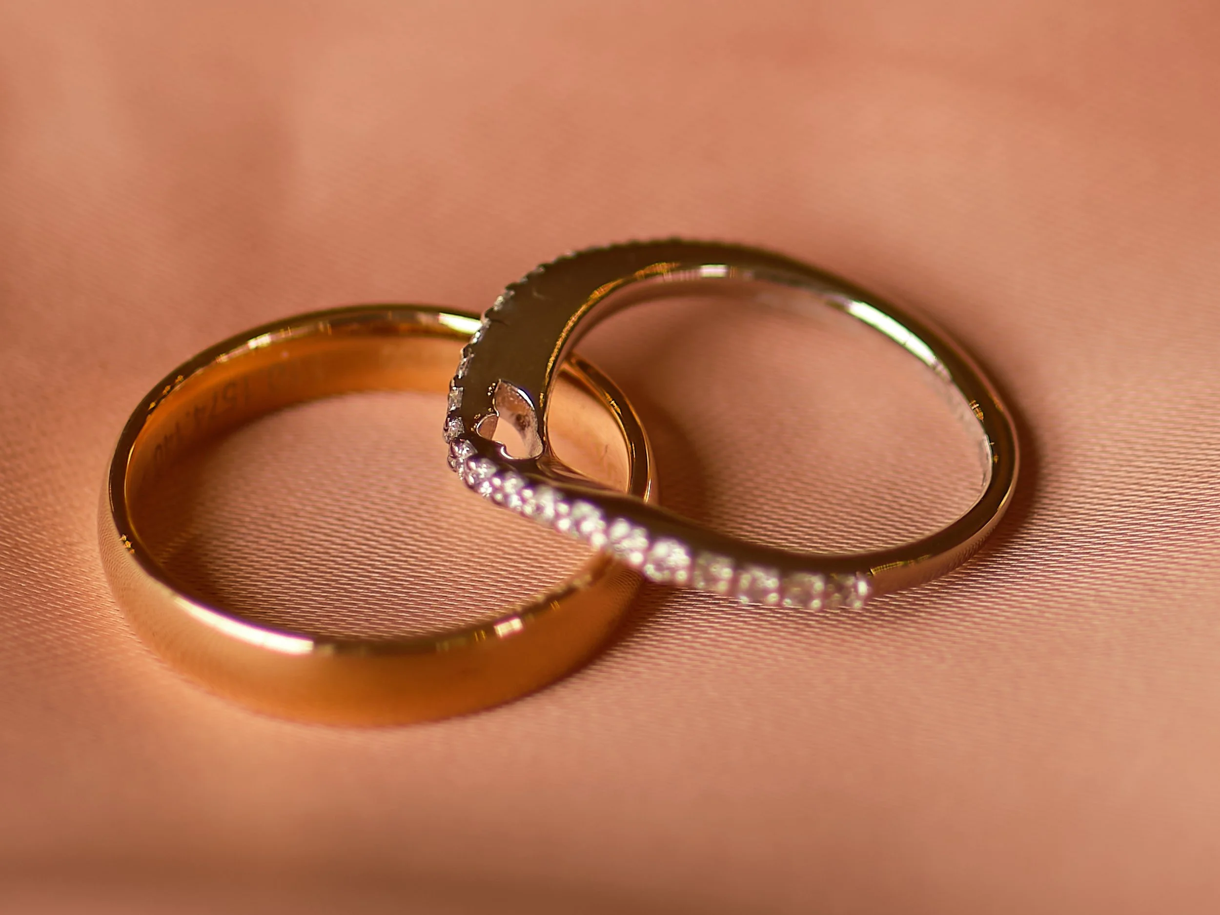 Two gold wedding bands, one plain and one with small diamonds, resting on pink fabric.