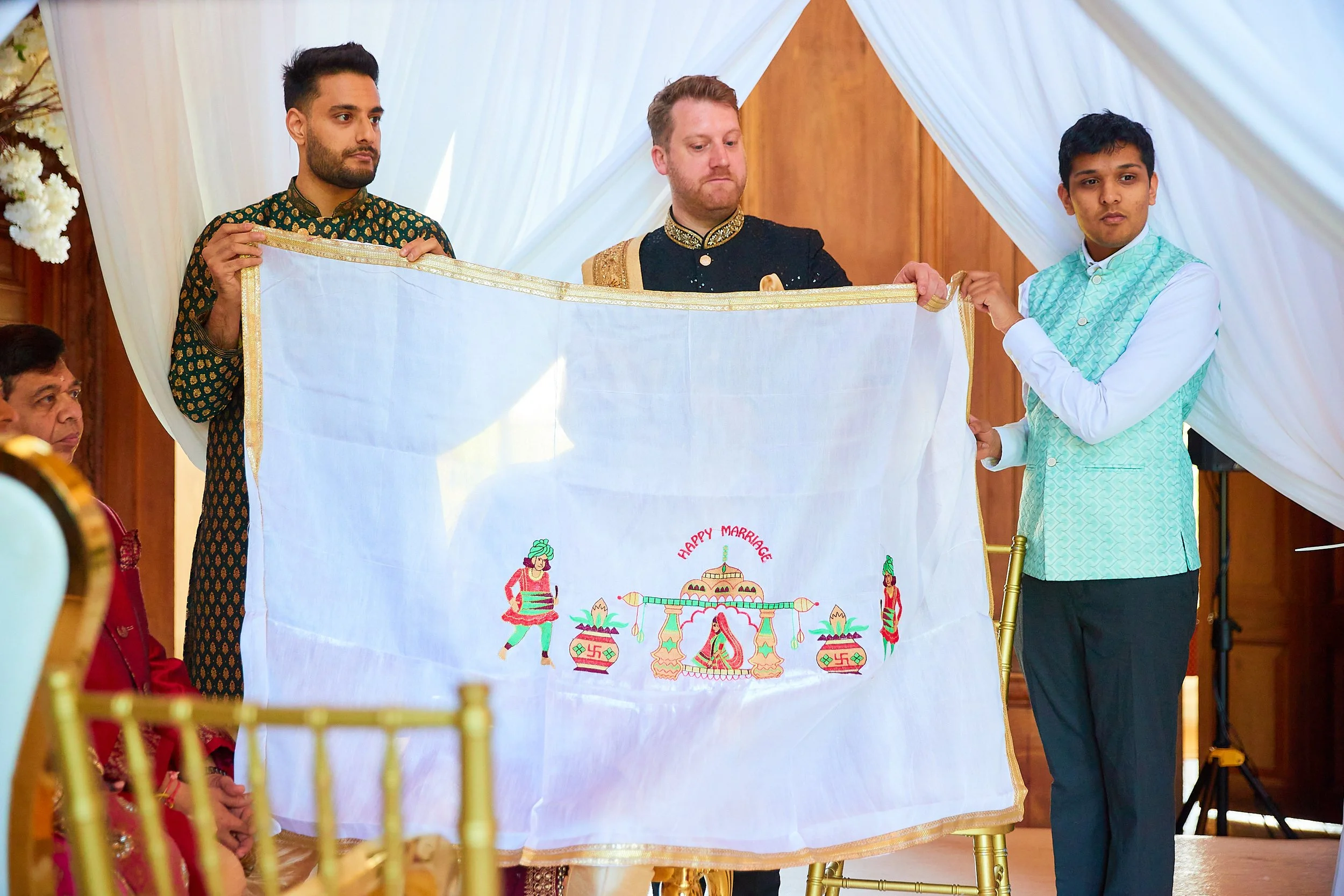 Three men hold a white cloth with embroidery during an Indian wedding ceremony. The cloth has images of traditional Indian dancers, a palace, and the words 'Happy Marriage' embroidered on it.