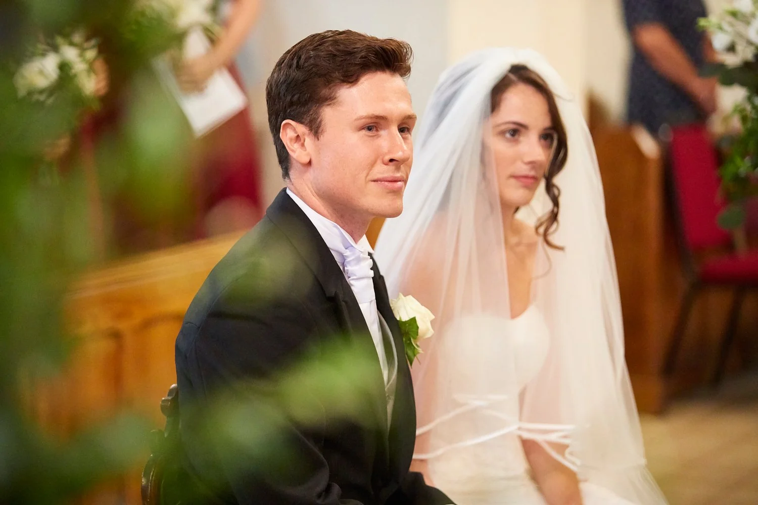 A groom and bride sit during their wedding ceremony inside a church, with the groom in a black tuxedo with a white boutonniere and the bride in a white wedding dress with a veil.