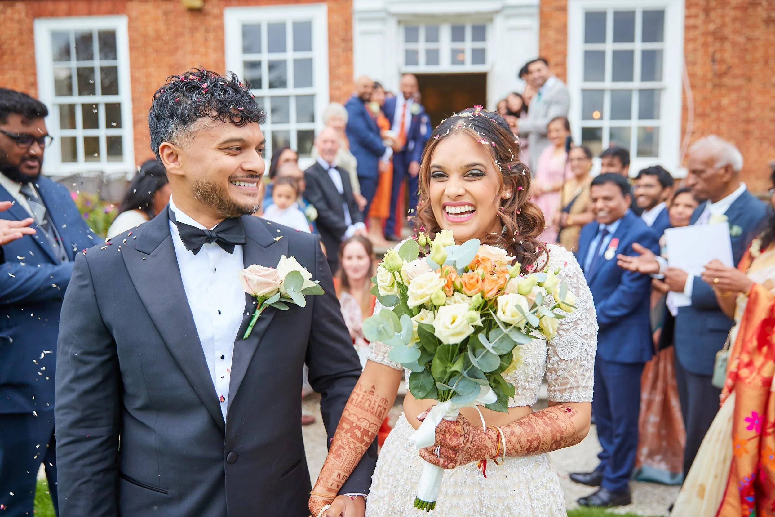 A happy bride holding a bouquet of flowers at her wedding, standing next to her smiling groom, surrounded by wedding guests outside a brick building.