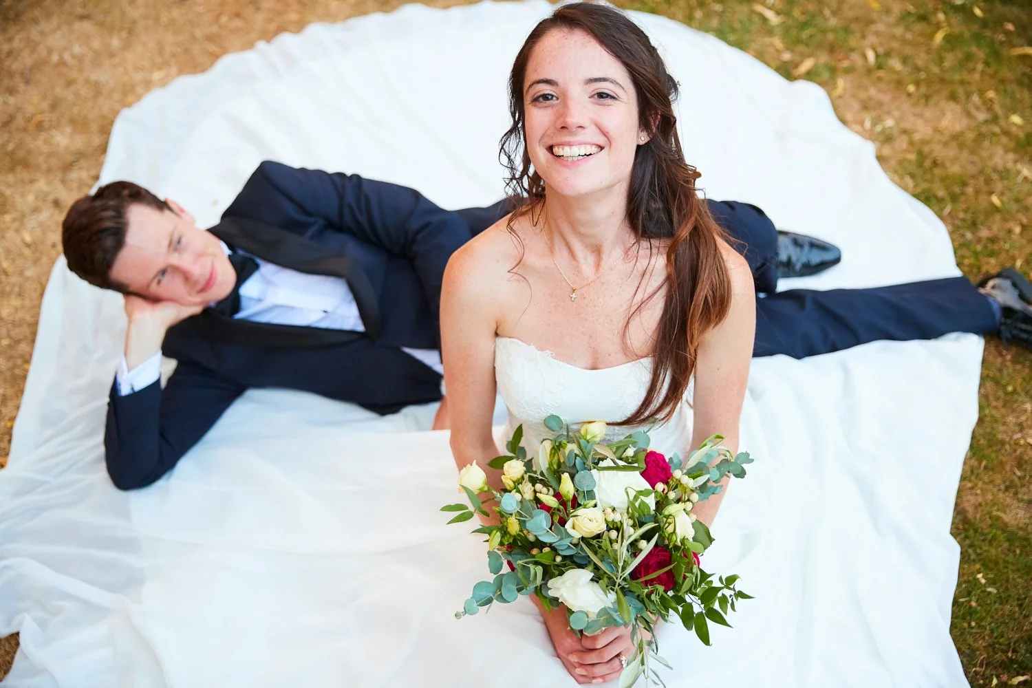 A bride holding a bouquet of flowers and a groom lying on the ground behind her on a white sheet, outdoors on grass.