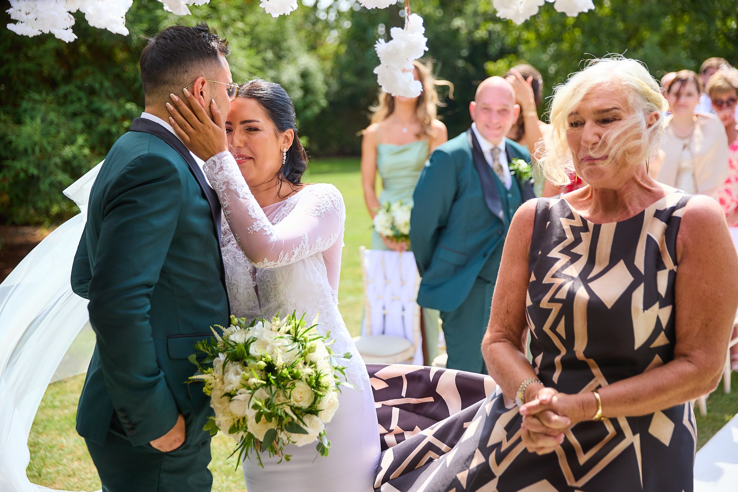 A bride and groom sharing an intimate moment at an outdoor wedding ceremony, with a woman in a patterned dress in the foreground and other guests smiling in the background.