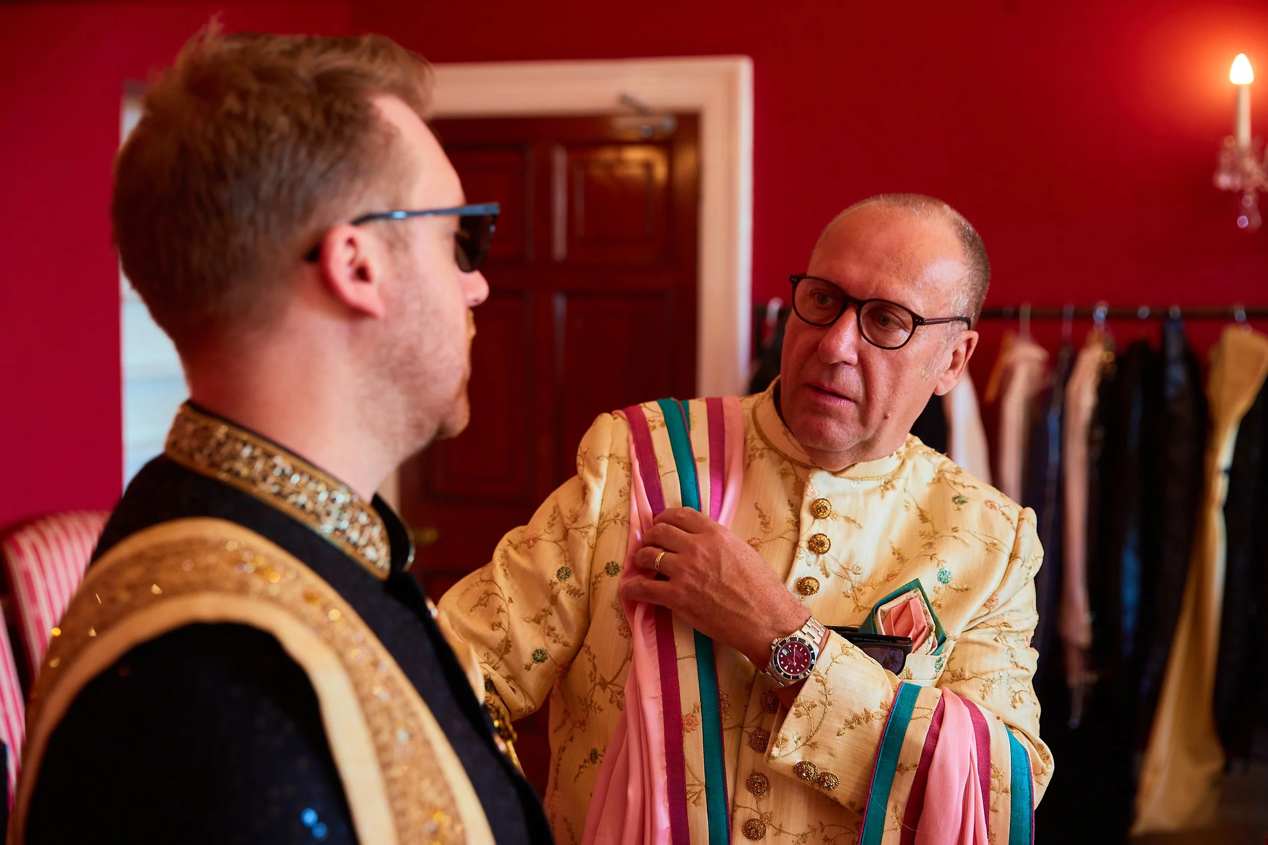 Two men dressed in ornate traditional clothing conversing indoors with a red wall and wardrobe behind them.