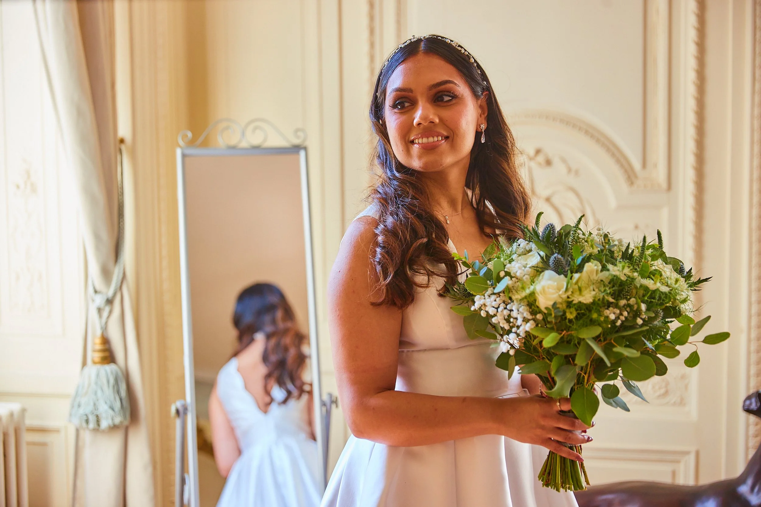 A bride with wavy brown hair, wearing a white dress and a headband, holding a bouquet of green and white flowers, standing in a decorated room with ornate walls and curtains.