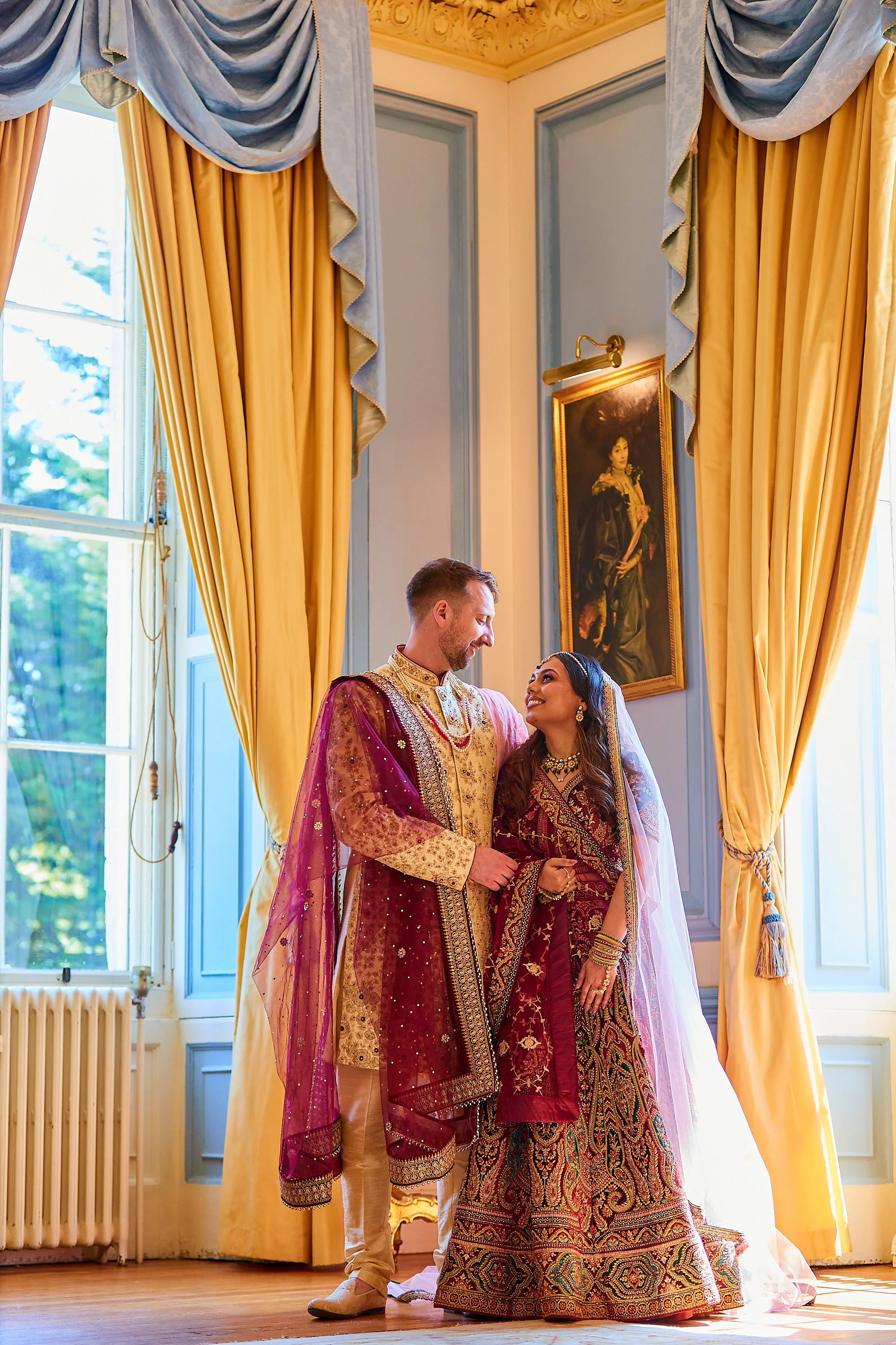 A couple dressed in traditional Indian wedding attire standing together in a sunlit room with large yellow curtains and a painting on the wall.
