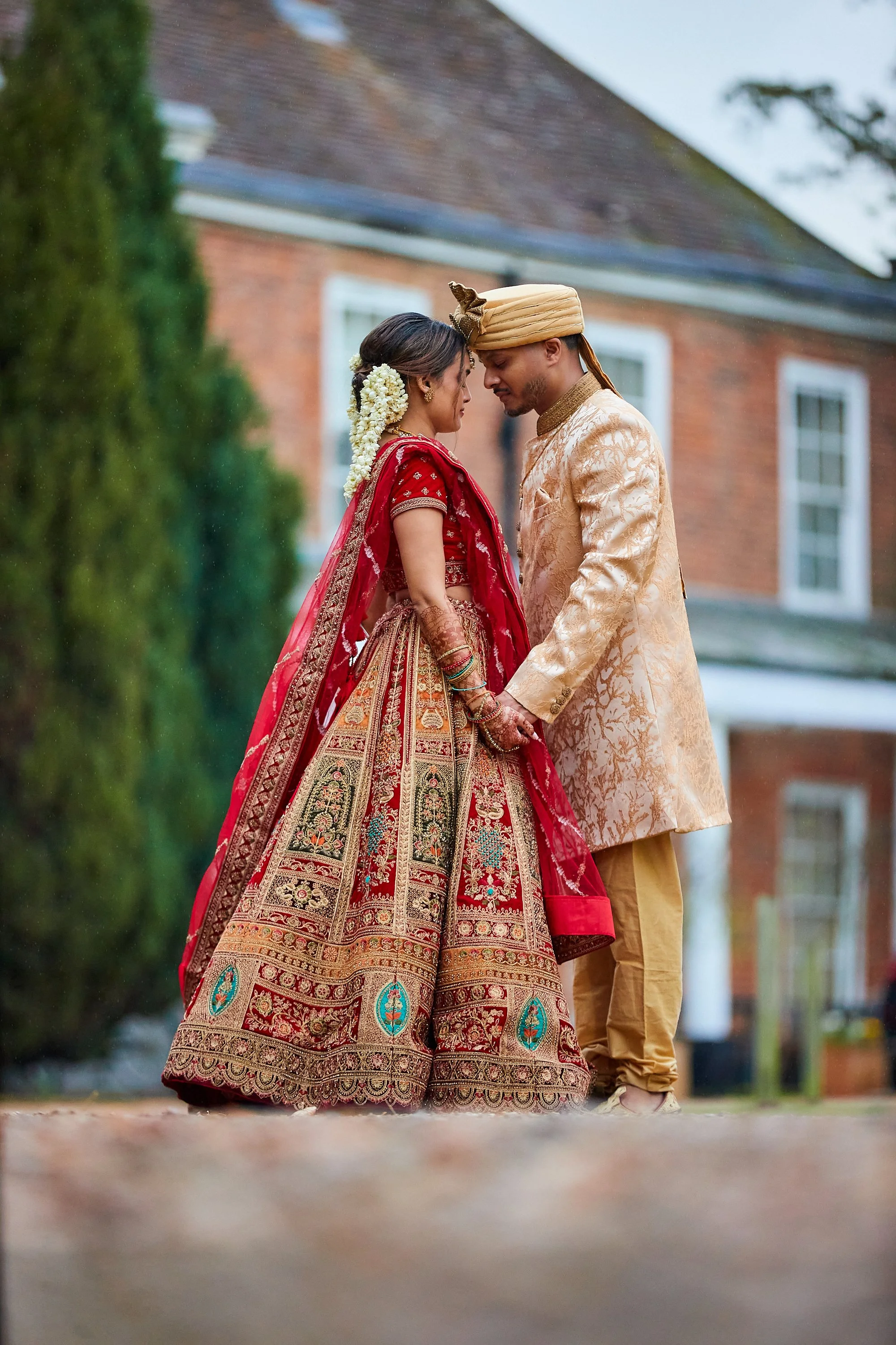 A couple in traditional Indian wedding attire standing close, touching foreheads, outdoors in front of a brick house.