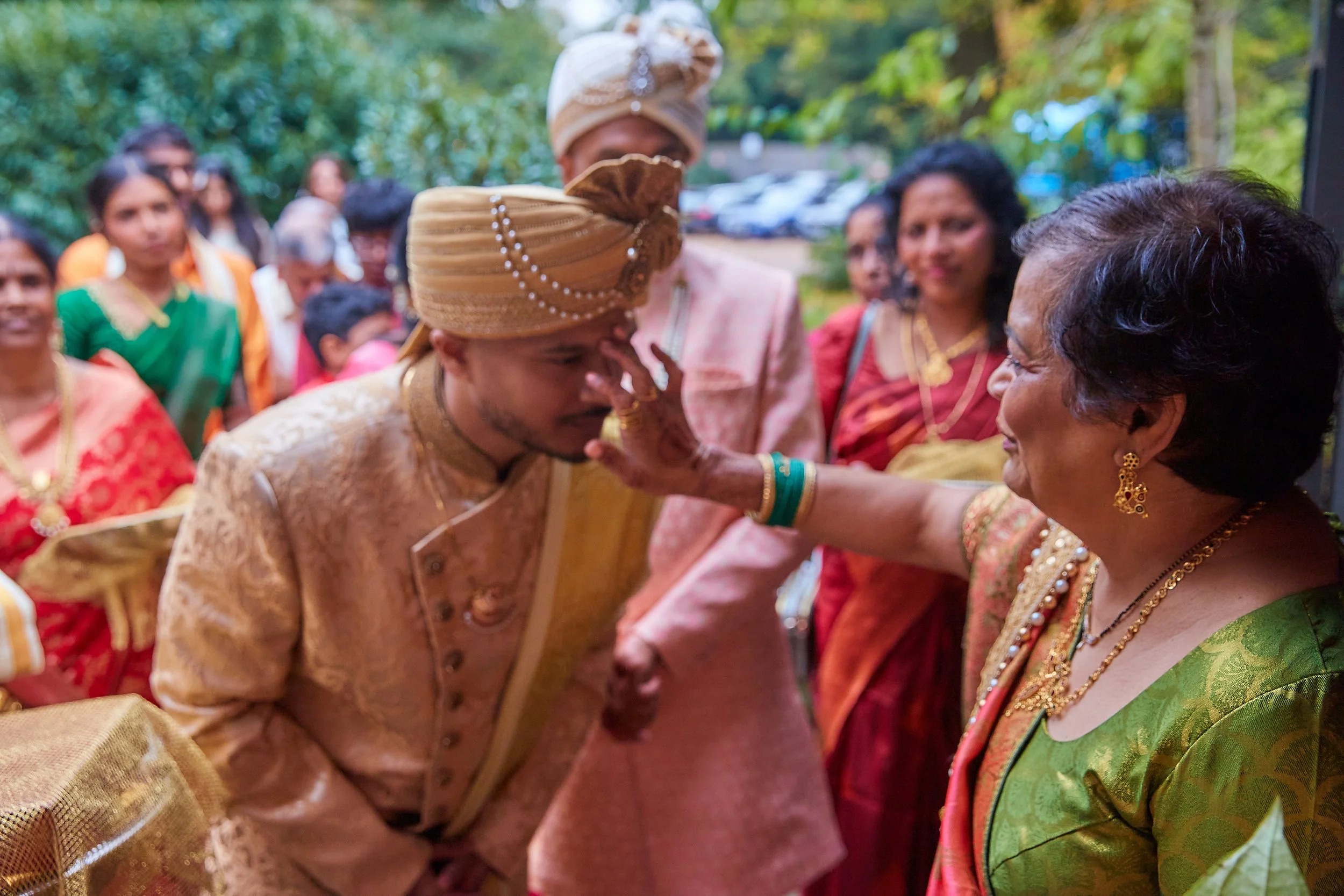 A wedding ceremony with Indian traditional attire, where a bride and groom are participating, and an elder woman is applying a ceremonial mark on the groom's forehead, surrounded by guests in colorful sarees and traditional clothing outdoors.