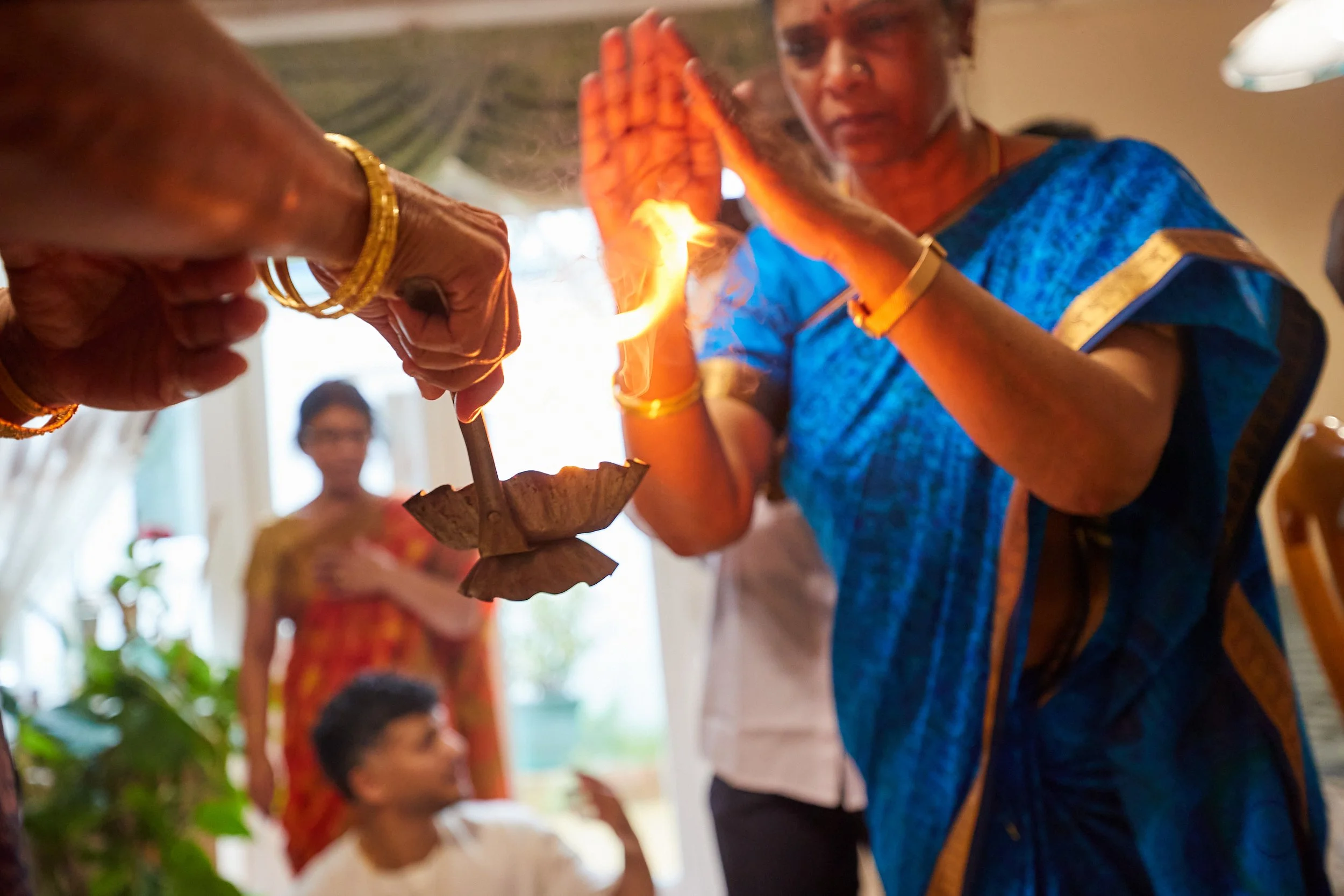 Women performing a traditional diya lamp lighting ceremony during a religious or cultural event, with a young girl and other people observing in the background.