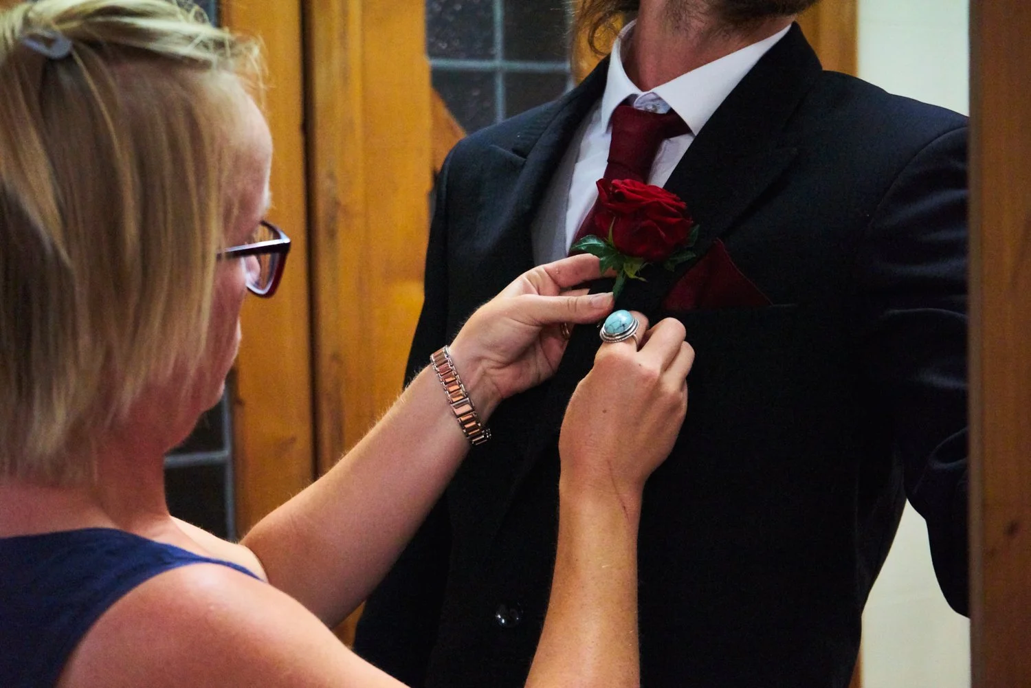 A woman pinning a red rose boutonniere onto a man's black suit jacket.