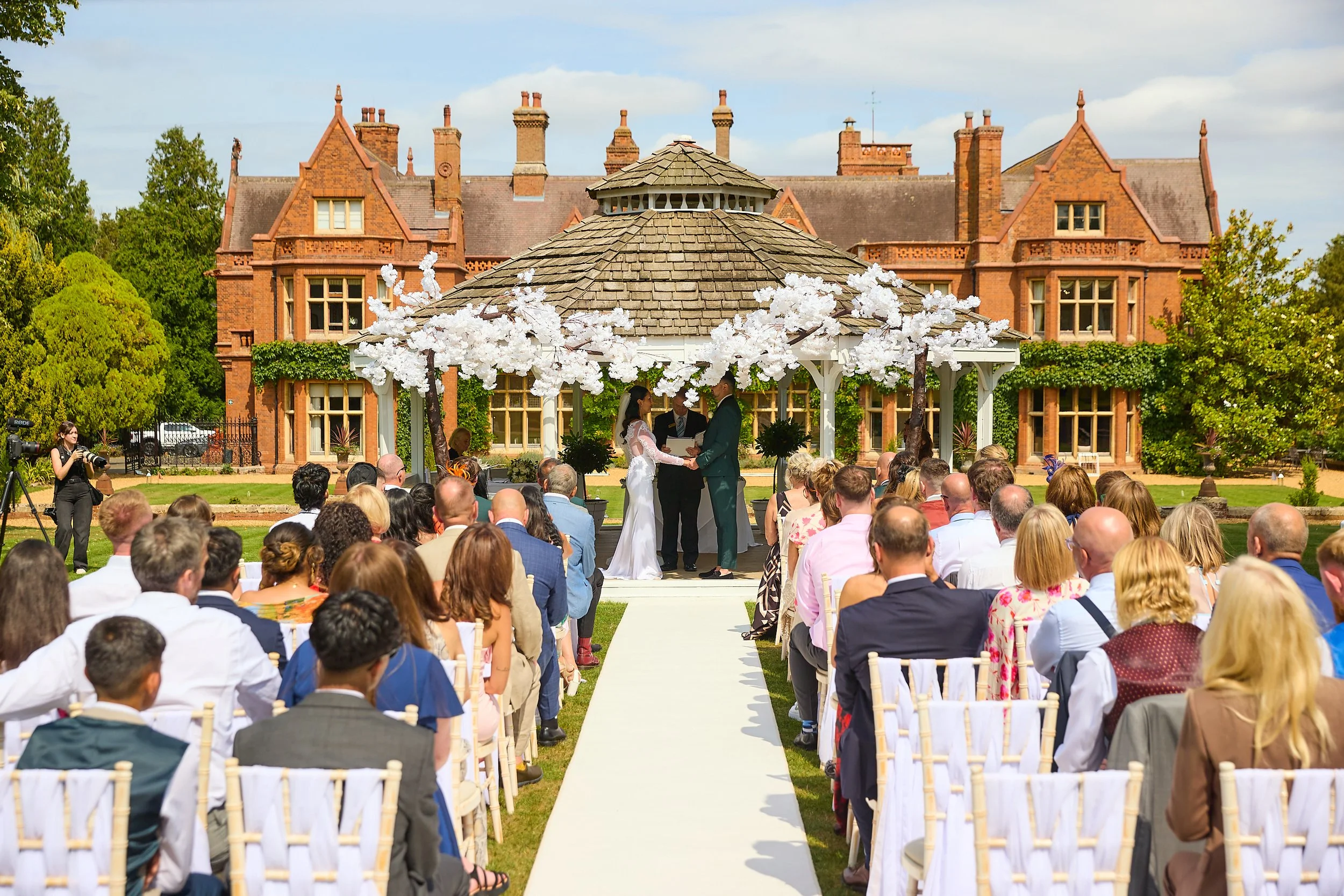 A wedding ceremony taking place outdoors on the lawn of a large historic brick mansion. The bride and groom are holding hands under a white wedding arch decorated with white tree-like structures, with guests seated facing them.