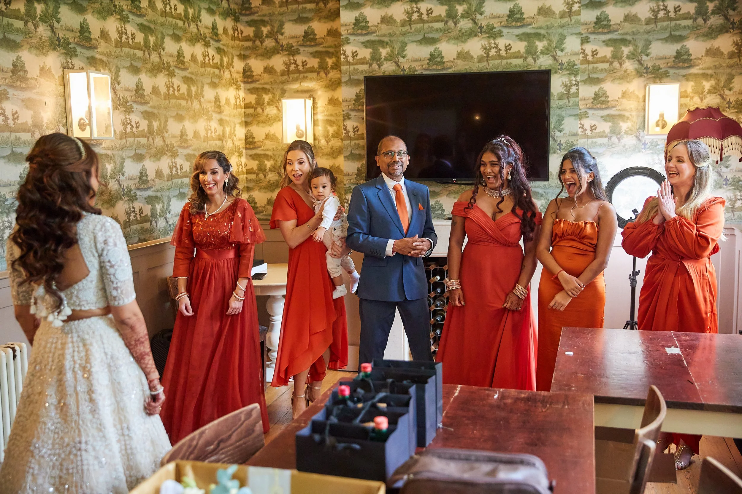 A group of women and a man are gathered in a room, smiling and looking at a woman in a white dress. The women are dressed in red and orange, and the man is in a blue suit with an orange tie. There is a child being held by one of the women. The room h