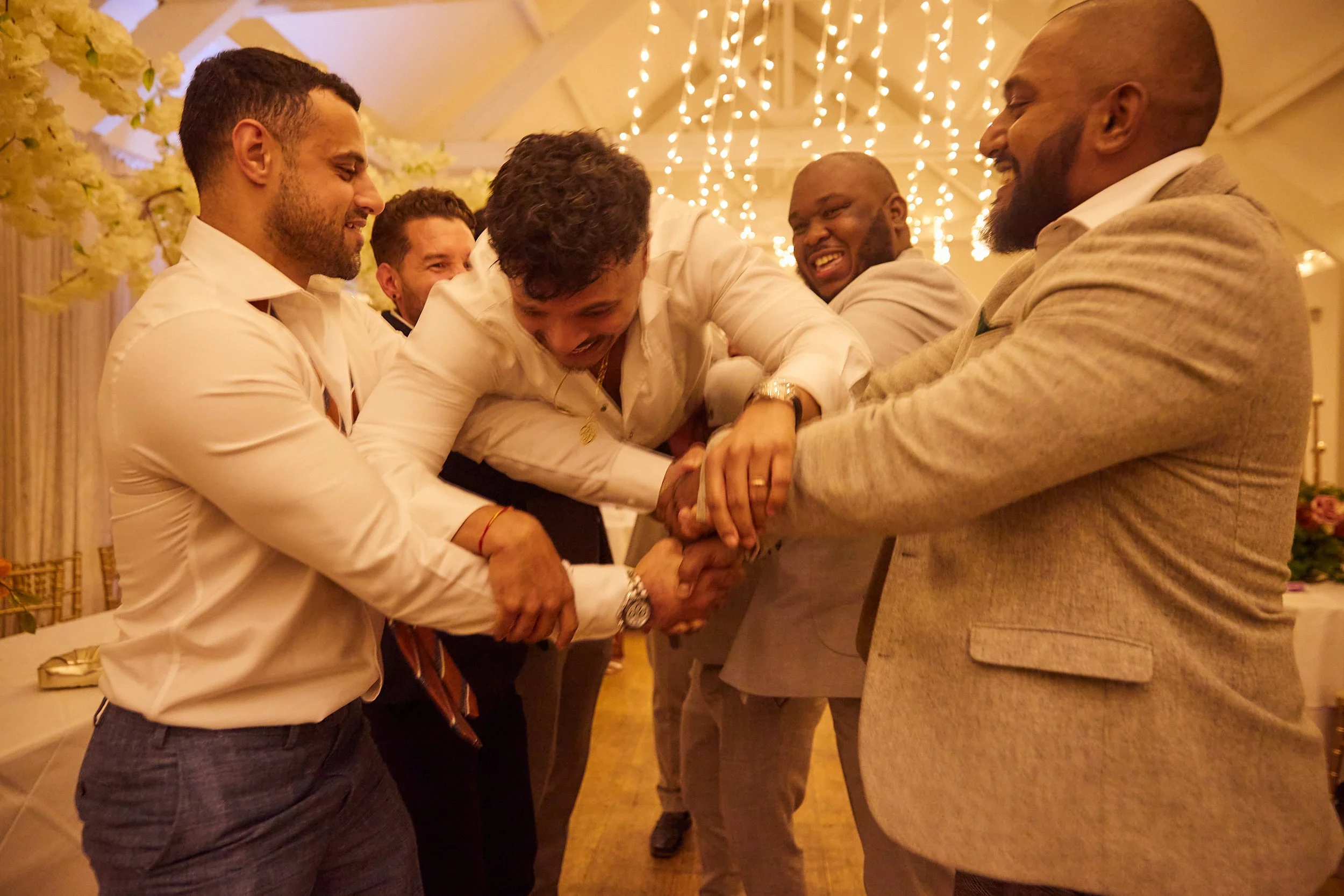 Group of men laughing and playfully holding each other at a celebration or wedding reception, decorated with string lights and floral arrangements.