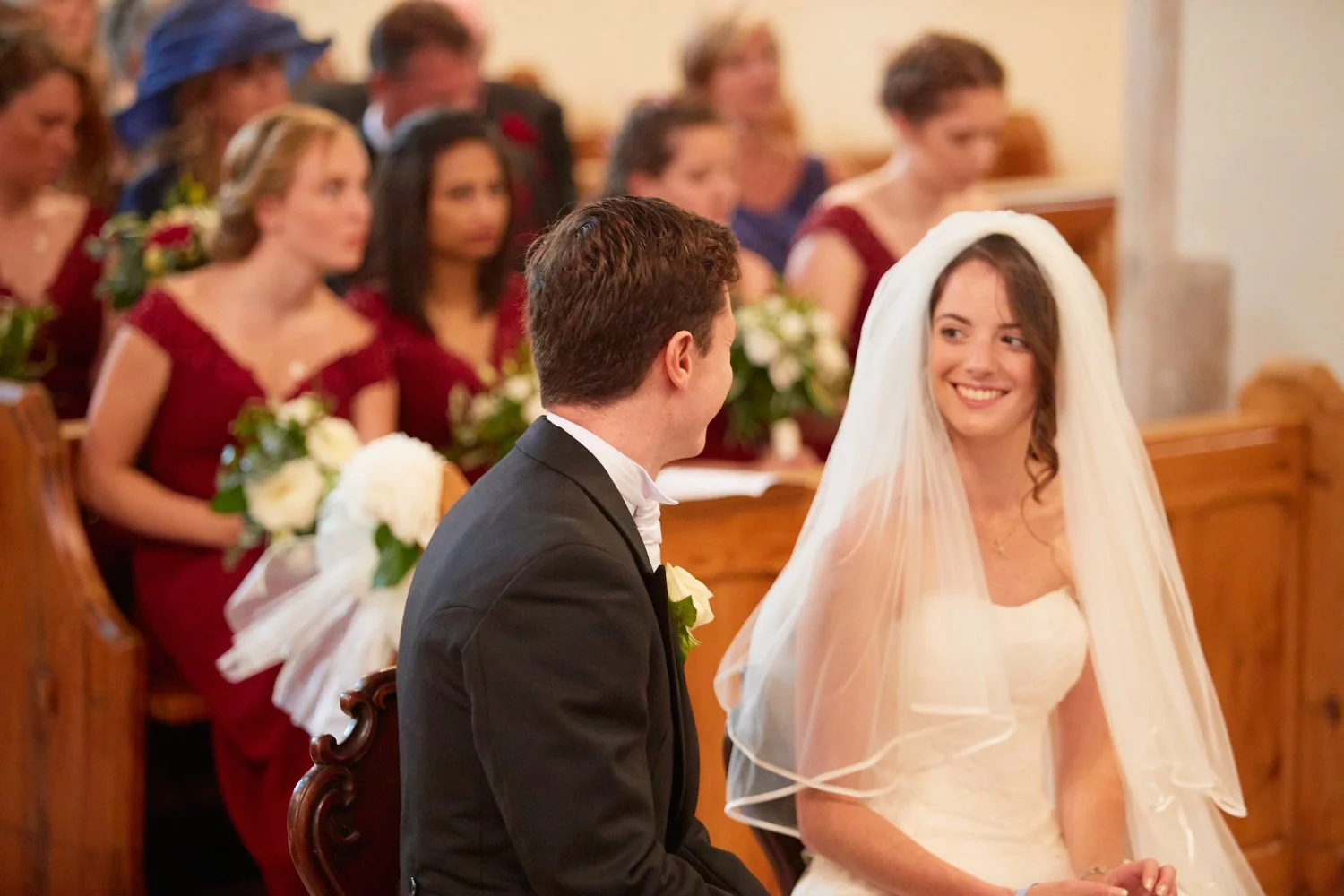 A wedding ceremony with a bride and groom sitting together, smiling at each other, in front of seated guests inside a church.