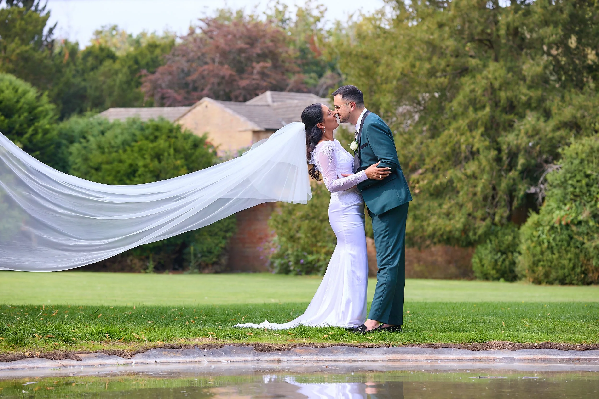 A bride in a white wedding dress and veil and a groom in a teal suit and glasses kiss outdoors on a grassy area with trees and houses in the background.