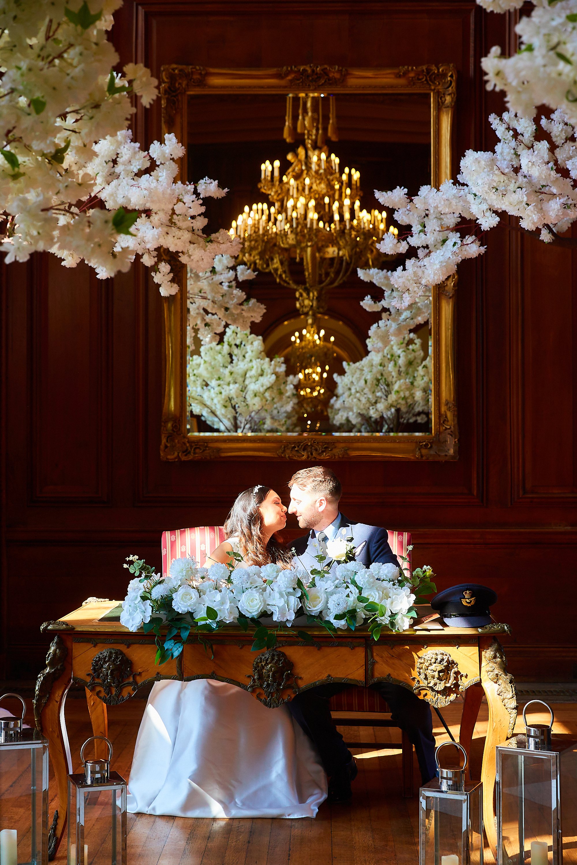 A newlywed couple sitting at a decorated table with white flowers, in a lavish room with wood-paneled walls, a large mirror, and a chandelier, sharing a moment close to each other.